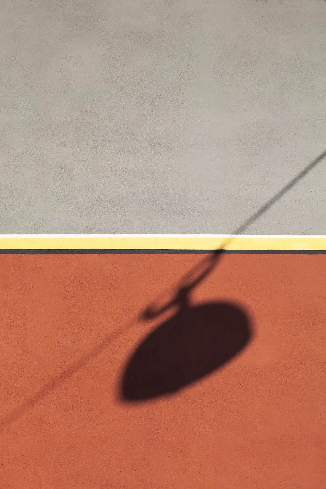 Ombra di una palla da tennis sul pavimento rosso e una parete bianca, con parte di un bastone o racchetta proiettata sulla parete
