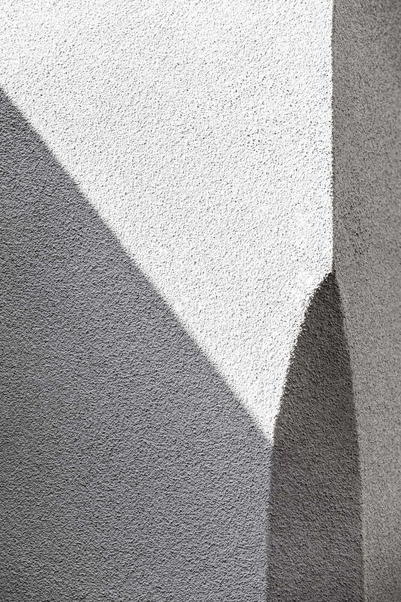Un muro bianco con una superficie ruvida, in piedi su un pavimento di cemento. La luce crea ombre sul muro, evidenziando la texture.
