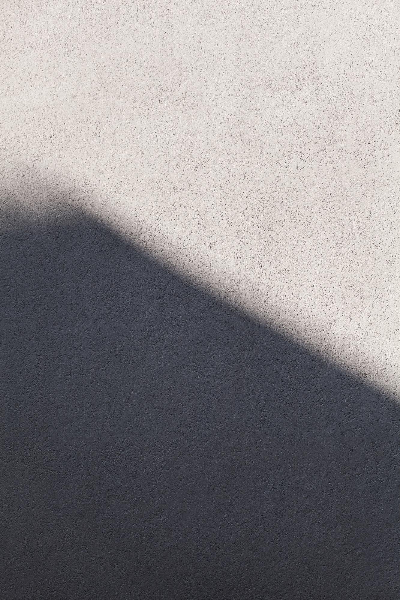 Superficie di muro con texture ruvida, metà superiore è chiara e metà inferiore è più scura, con un'ombra diagonale che divide le due sezioni.
