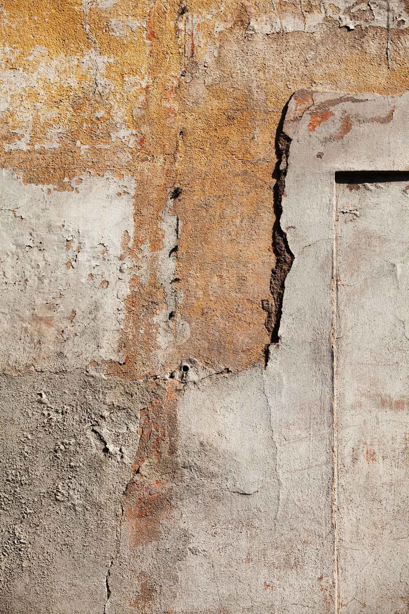 Prima di due foto di una parete di cemento con vecchie vernici e crepe, evidenziando lo stato di deterioramento dell'edificio.