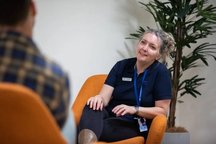 A woman with gray hair, wearing a navy uniform and a lanyard, sitting in an orange chair, talking to a person in a plaid shirt. There is a tall plant behind her.