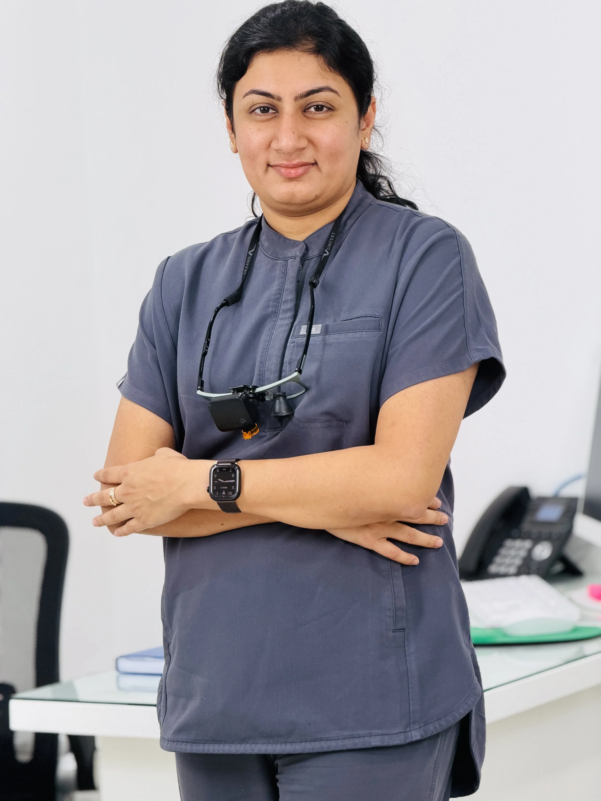 A woman in medical scrubs standing in an office with her arms crossed, wearing a smartwatch and a headlamp around her neck, with office furniture and a phone on a desk in the background.