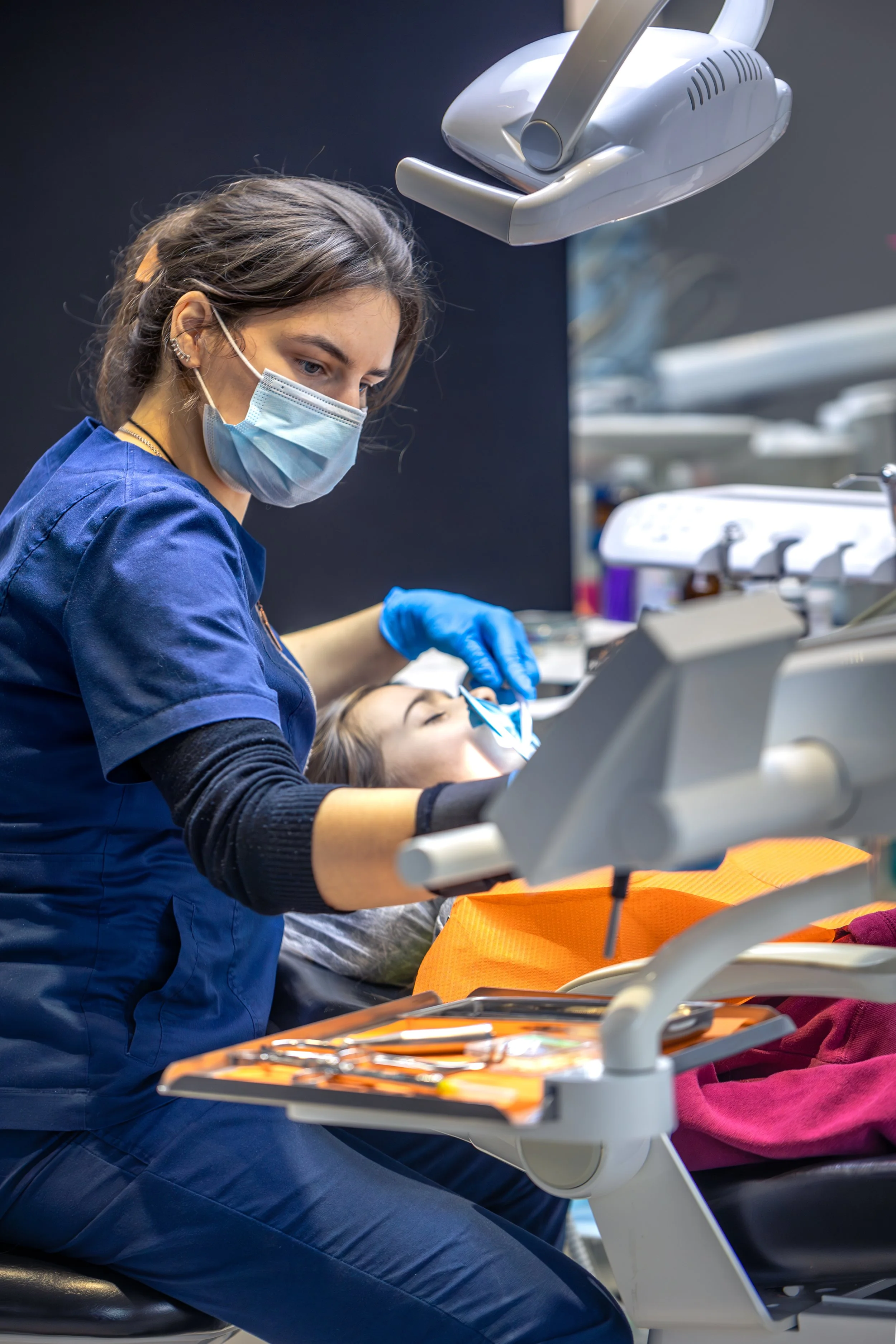 A dental professional in blue scrubs, wearing a face mask and gloves, attended by a patient lying in a dental chair. The professional is working on the patient's mouth with dental tools, with dental equipment above and around them.