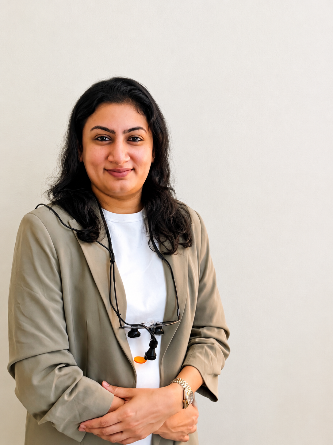 A woman with dark hair and a beige blazer wearing a white shirt with magnifying glasses around her neck, standing against a plain off-white wall.