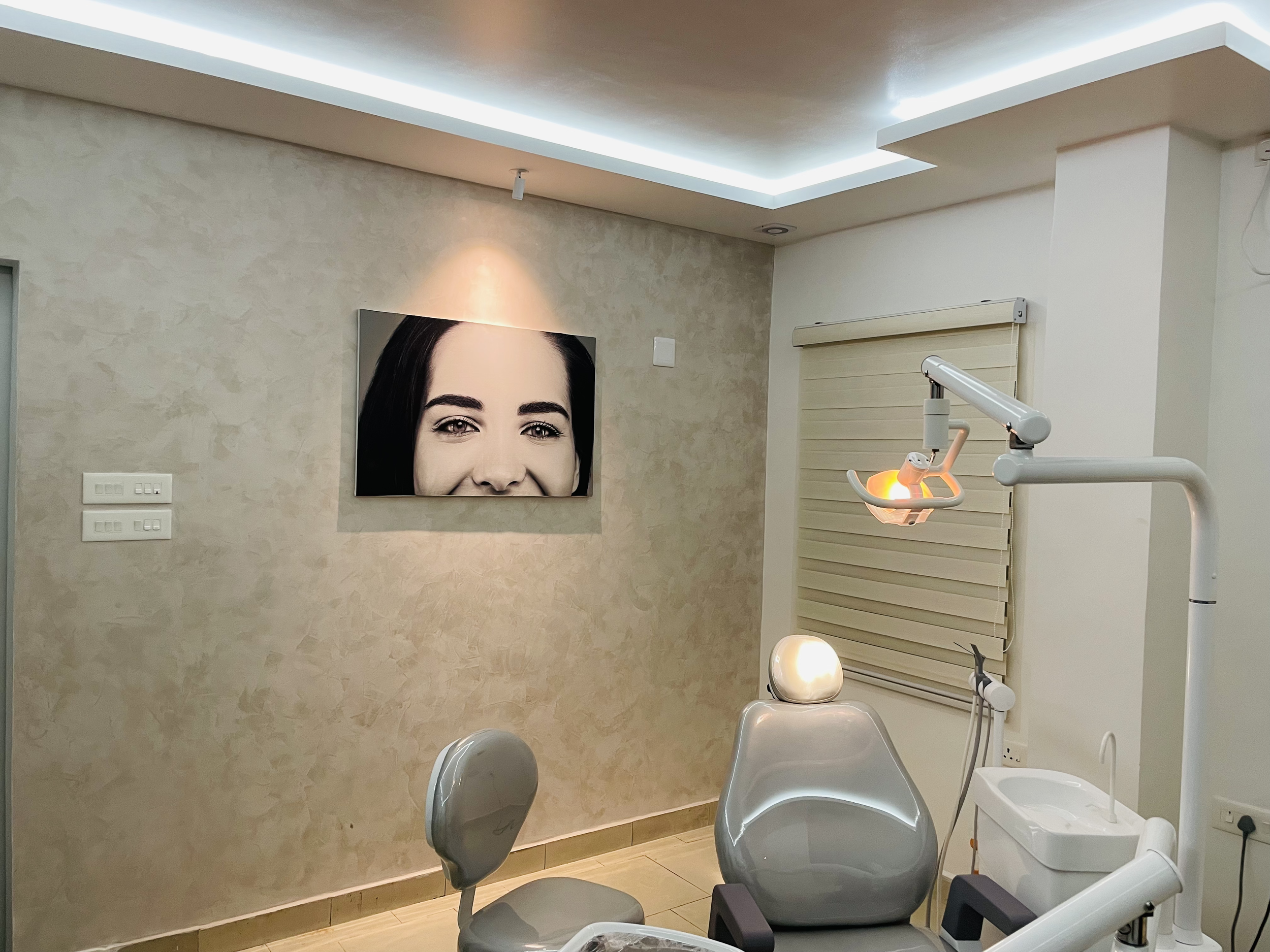A dental examination room with dental chairs, a dental lamp, a portrait of a smiling woman on the wall, and utility panels on the wall.