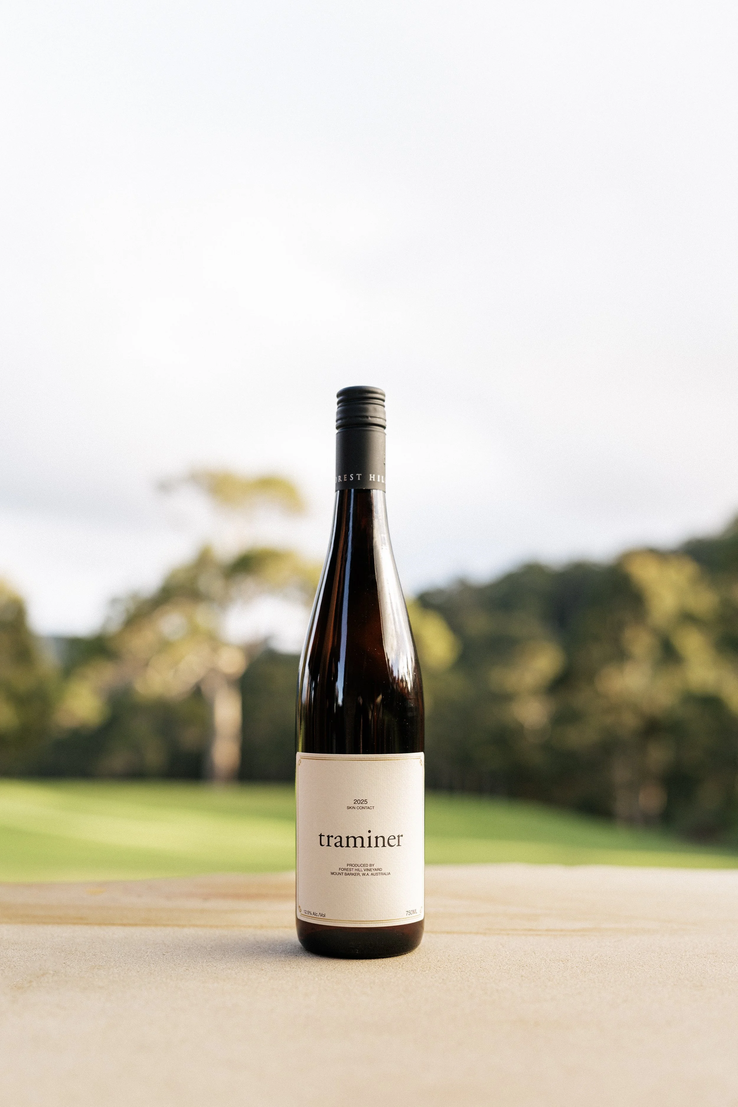 A wine bottle with a white label stands on a surface outdoors with a background of green grass and trees, under a cloudy sky.