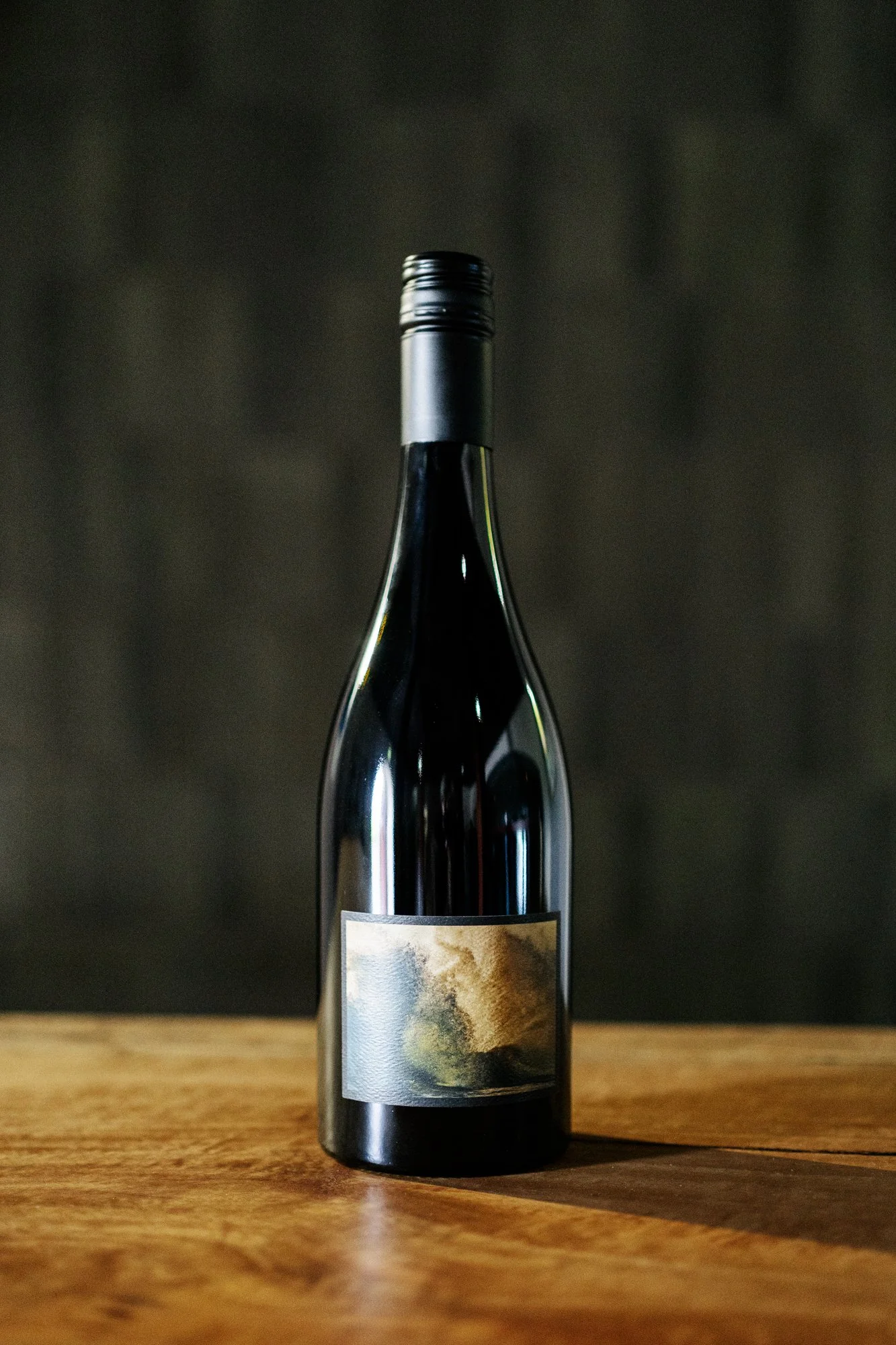 A dark-colored wine bottle with a screw cap and a landscape painting on the label, placed on a wooden surface with a dark background.