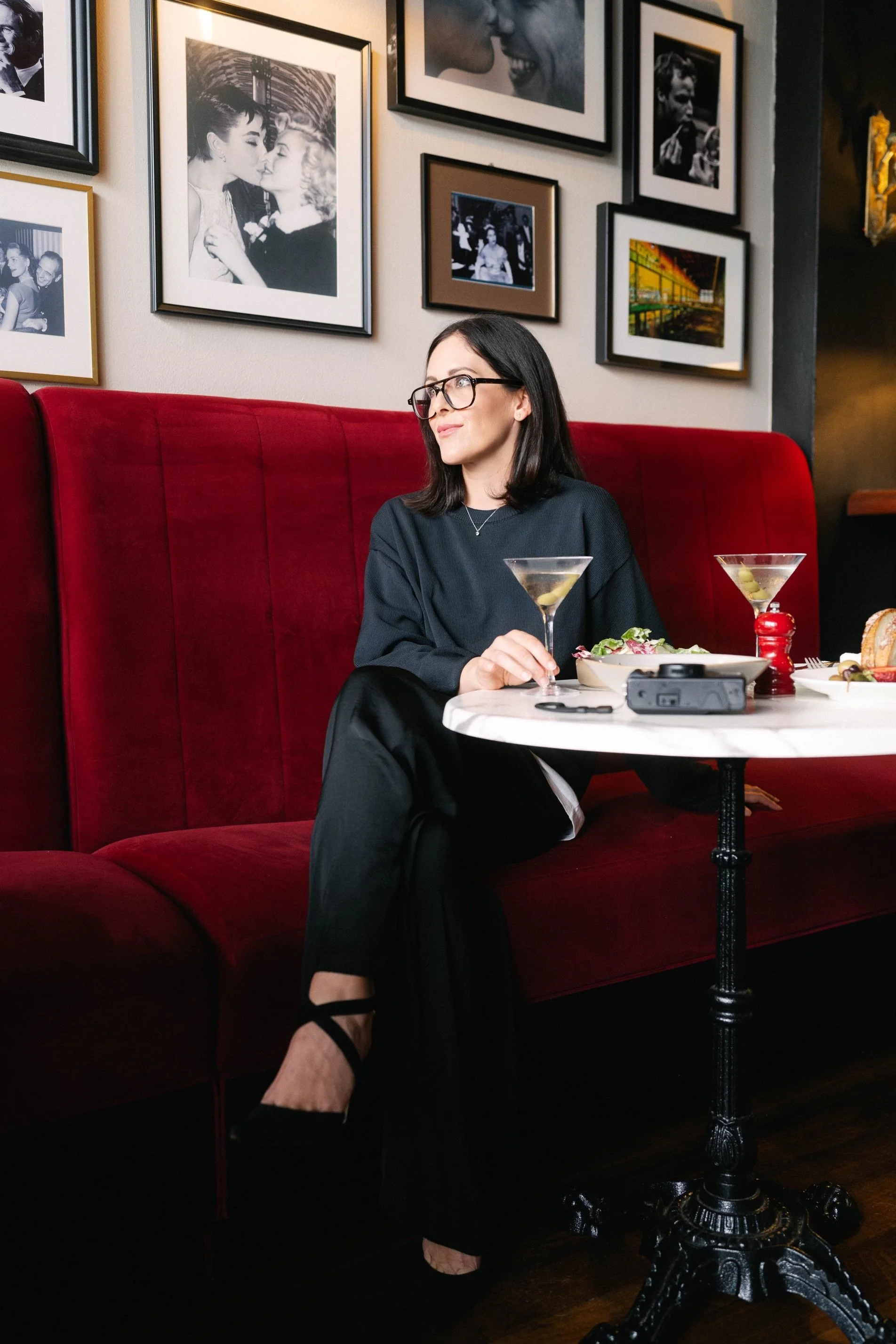 A woman with black hair and glasses sits at a white marble table in a restaurant with red velvet seating. She is holding a martini glass and looking to her right. On the table are two martinis, a bowl of salad, a pepper shaker, and a camera. The wall behind her features a gallery of black-and-white and colorful framed photographs of people and cityscapes.