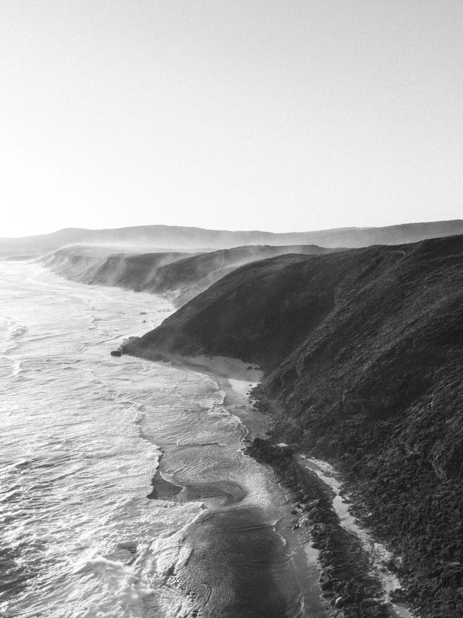 Black and white coastal landscape with cliffs, waves crashing on the shore, and hills in the background under a clear sky.