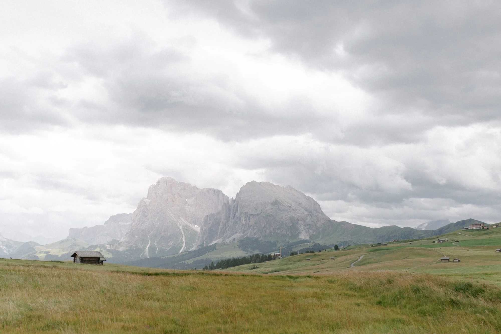 Scenic landscape of mountains under cloudy sky with green meadows and scattered small buildings in the foreground.
