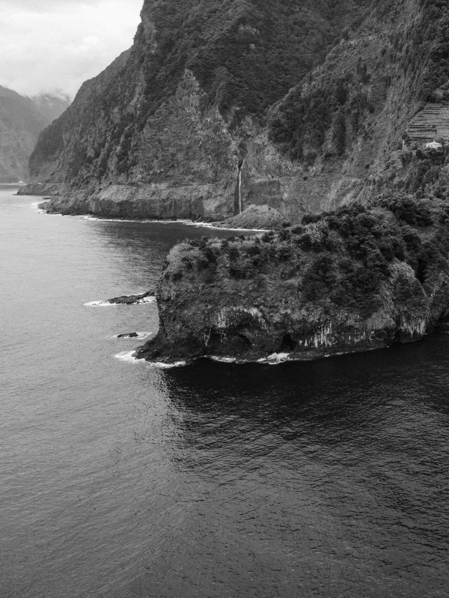 Black and white photo of a cliffside with a narrow waterfall, overlooking a body of water with gentle waves.