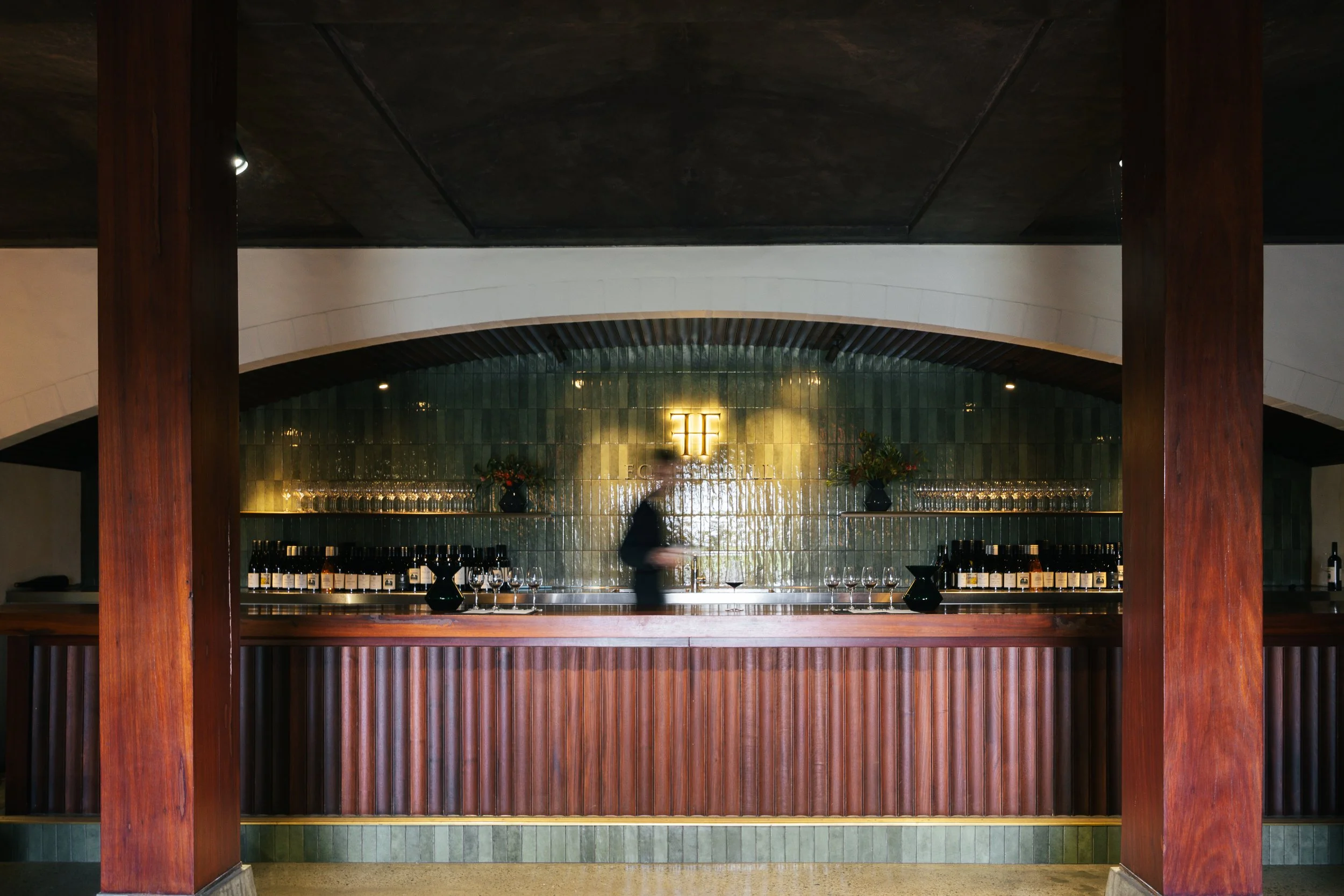A bar area with a polished wood counter, dark green tile backsplash, and a logo with the letters 'EF' illuminated behind the bar. There are wine glasses and bottles on the counter and two vases with flowers on the back shelf.