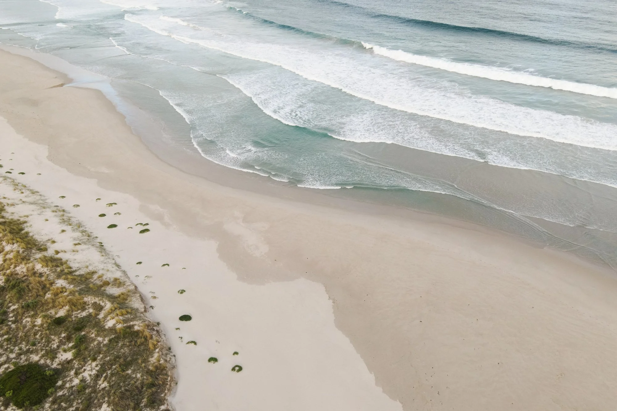 Aerial view of a beach with white sand, green shrubbery, and gentle ocean waves