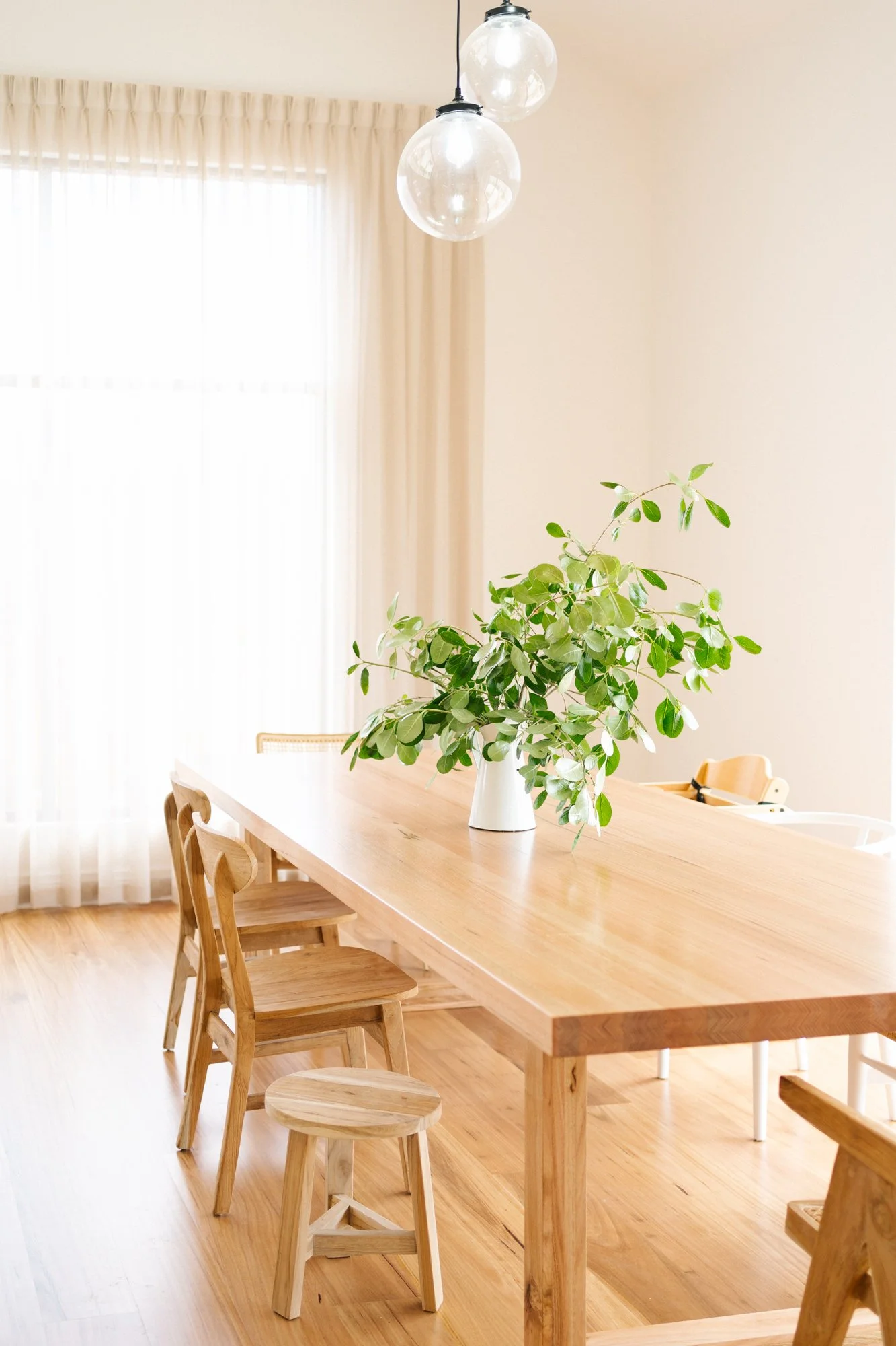 A wooden dining table with a white vase holding green leafy branches, surrounded by wooden chairs in a bright room with sheer curtains and hanging globe lights.