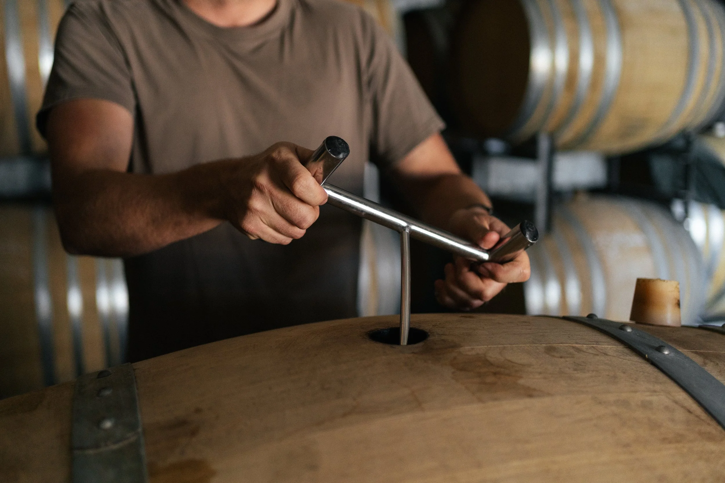 A person is holding a metal ring with one hand and inserting it into a hole in a wooden surface with the other hand, in a woodworking or craftsmanship setting, with barrels in the background.
