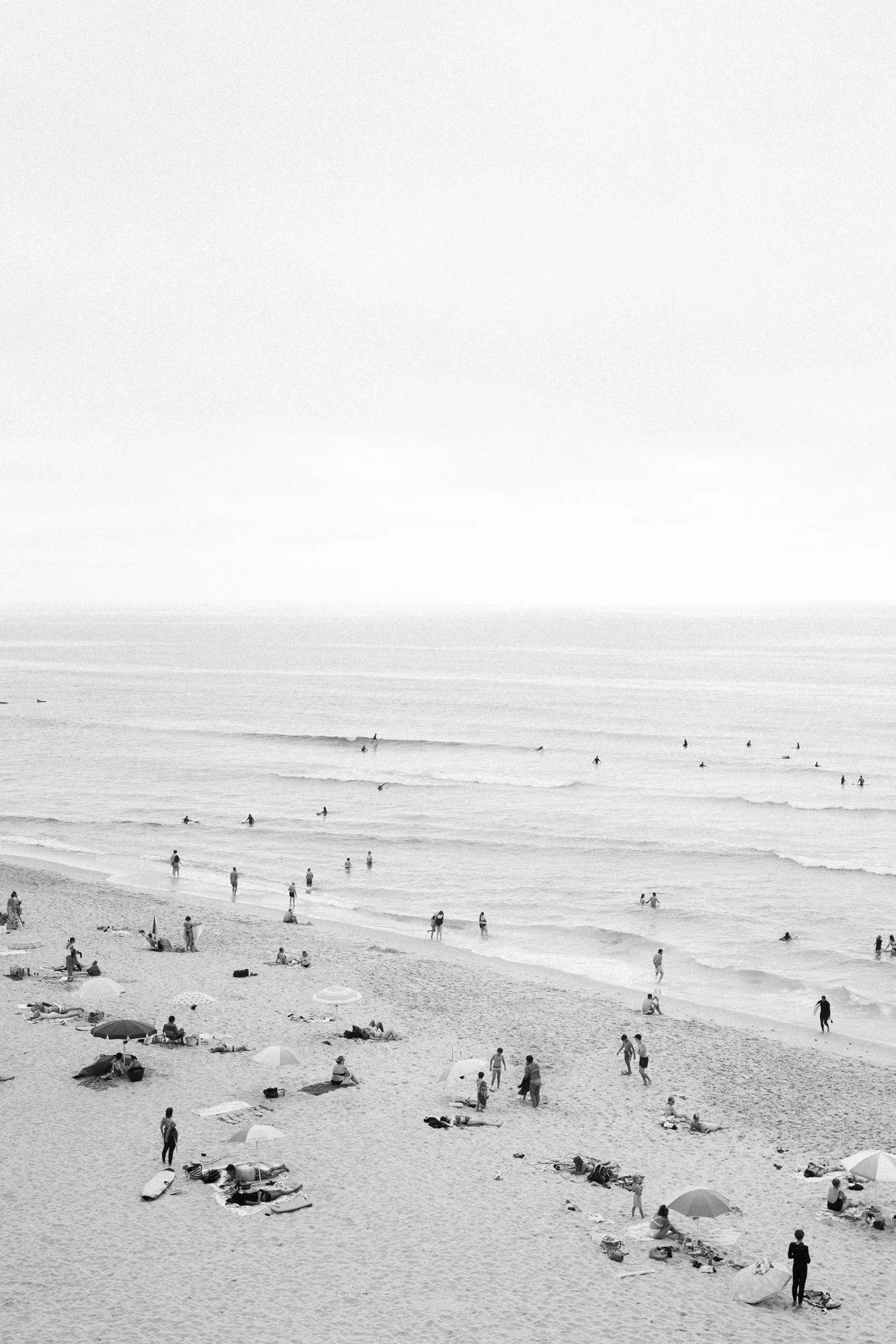 A black and white photograph of a crowded beach with people swimming, sunbathing, and walking on the sand. Beach umbrellas are scattered across the sandy shore, and the ocean waves are visible in the background.