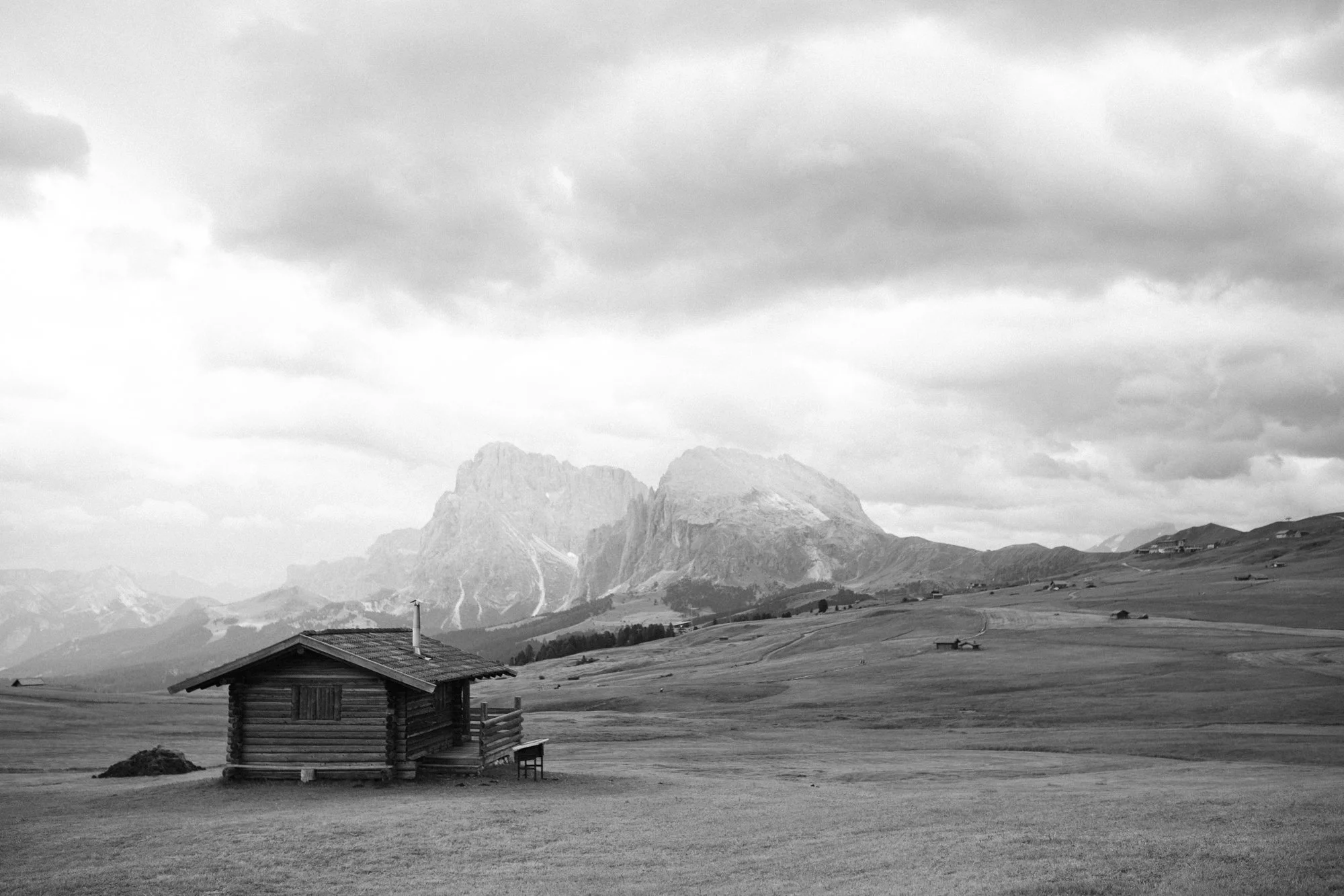 Black and white photo of a mountain landscape with a cloudy sky. There is a small wooden cabin in the foreground, surrounded by grassy fields and a few other small structures in the distance.