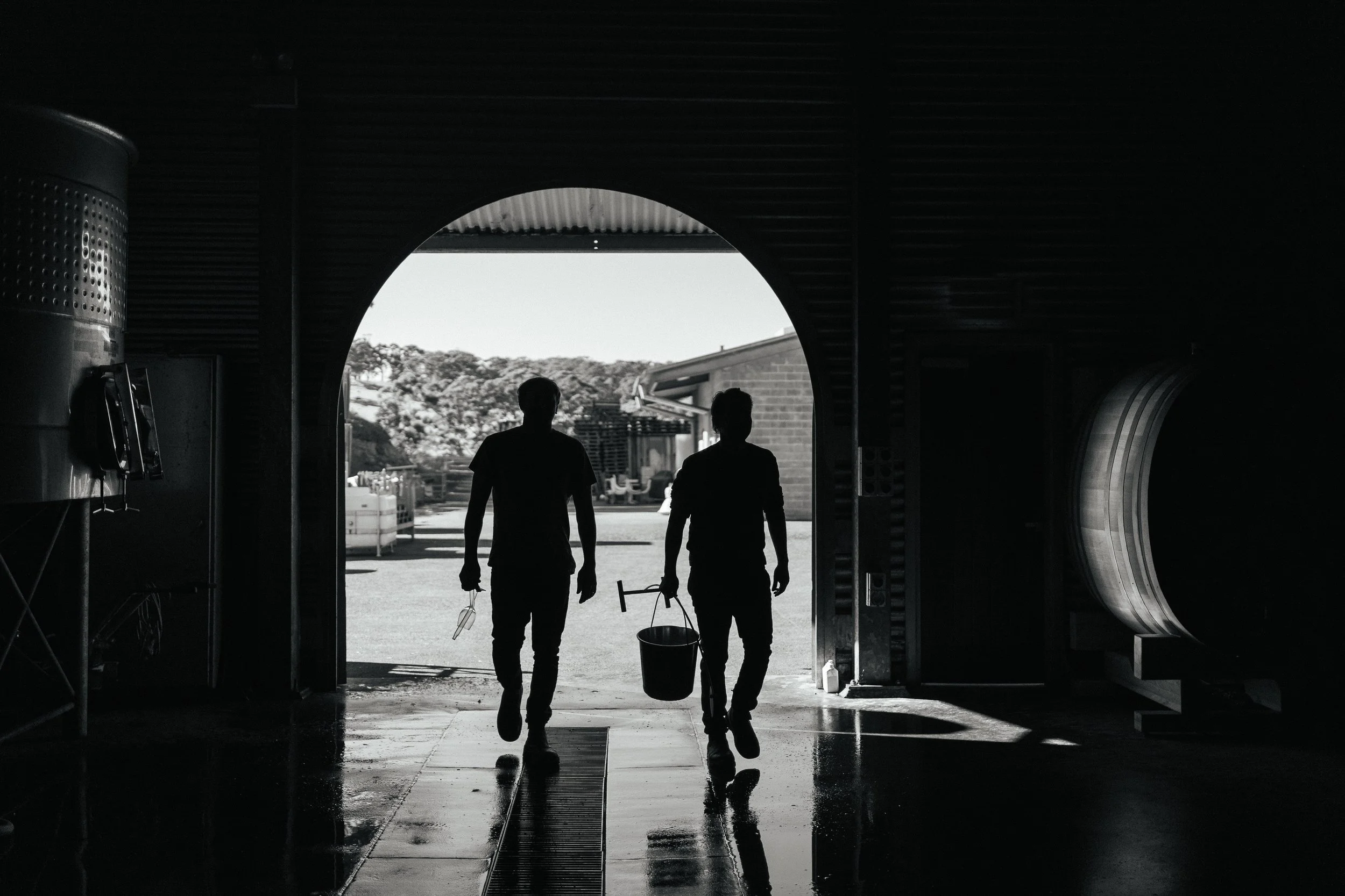 Two people silhouetted against the daylight walk out of a building, carrying buckets and tools, with an outdoor scene and trees in the background.