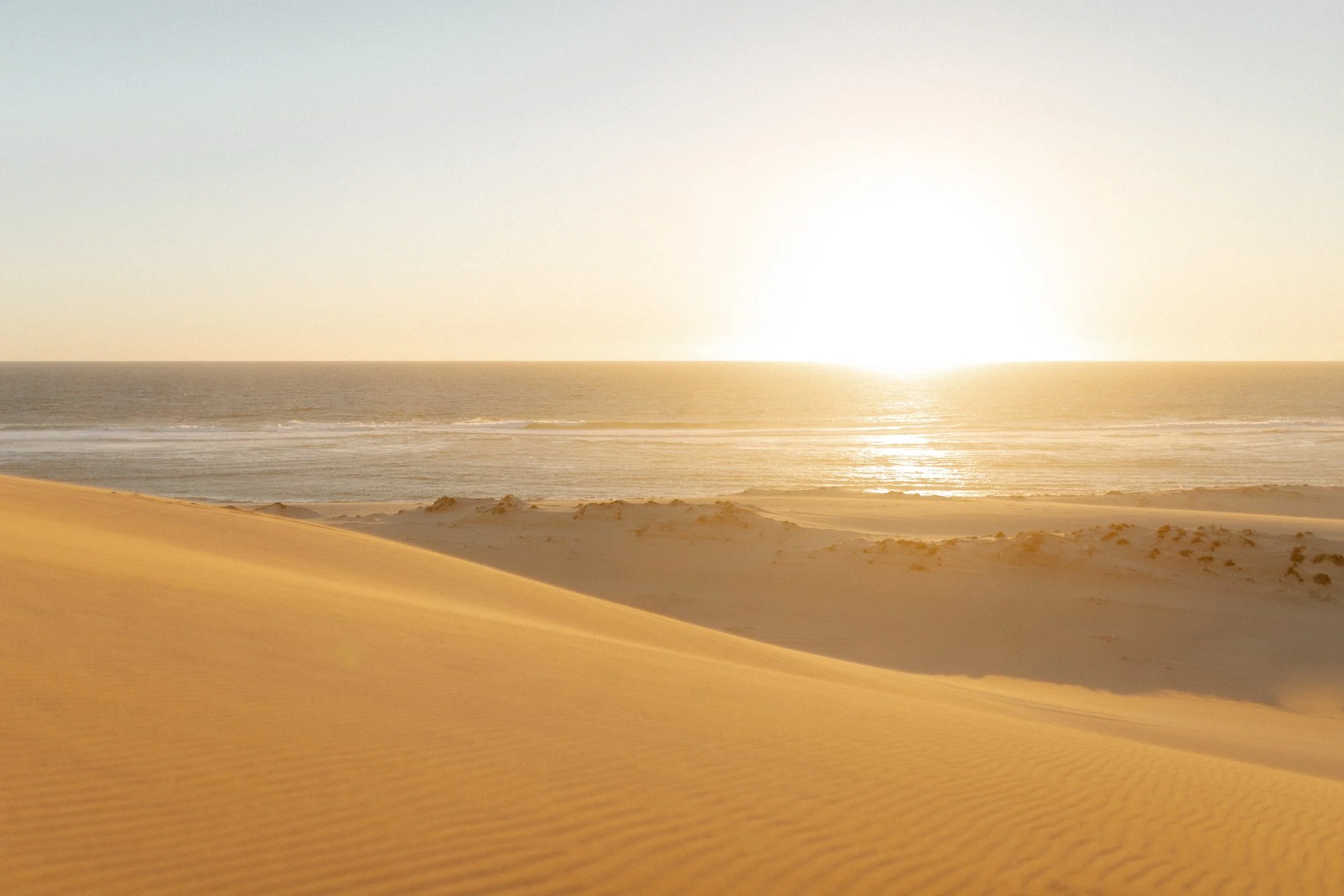 Beach with sand dunes, calm ocean waves, and the sun setting on the horizon.