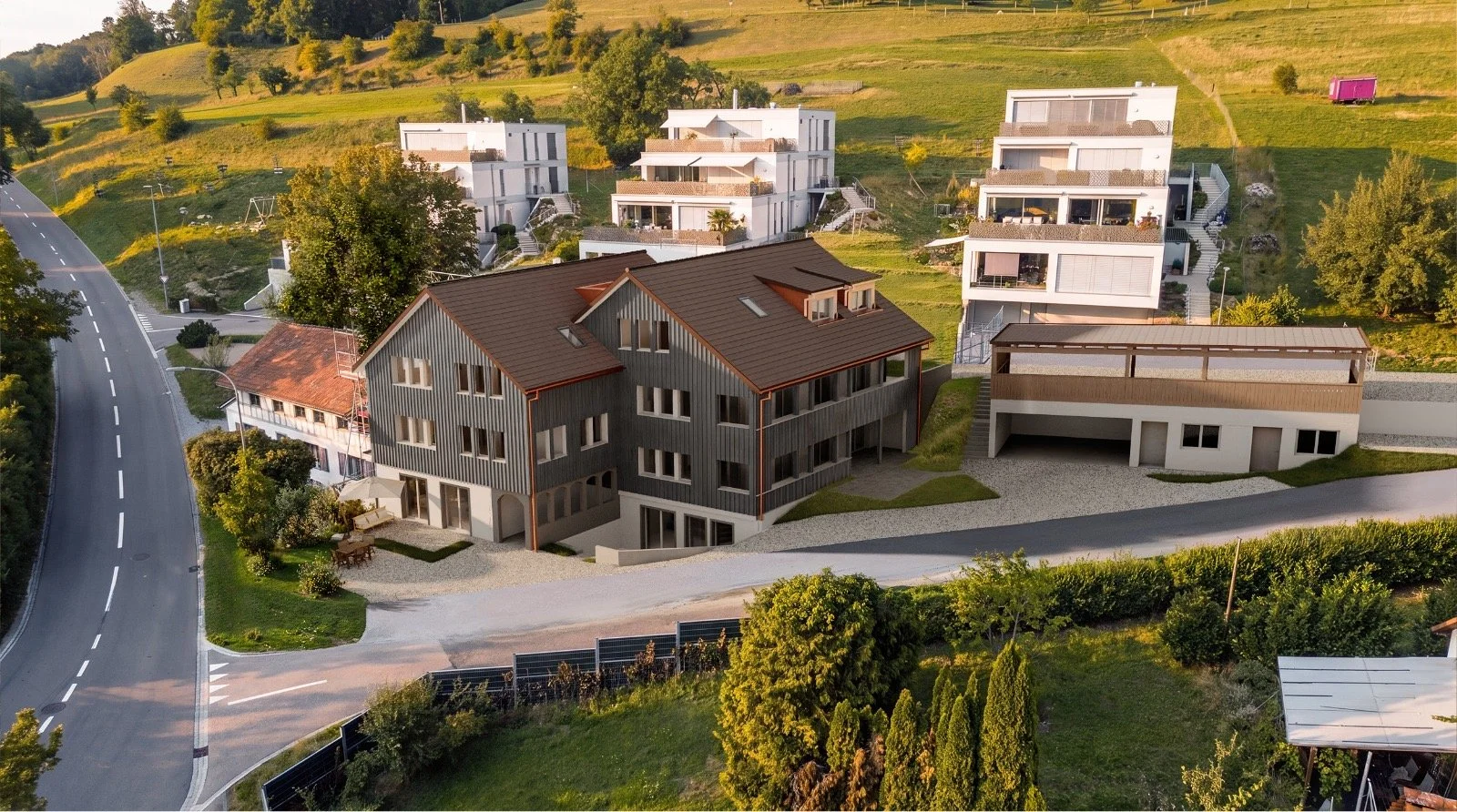 Mehrfamilienhäuser und Wohngebäude auf einer hügeligen Landschaft mit Bäumen und Straßen in der Nähe, fotografiert bei Sonnenuntergang.