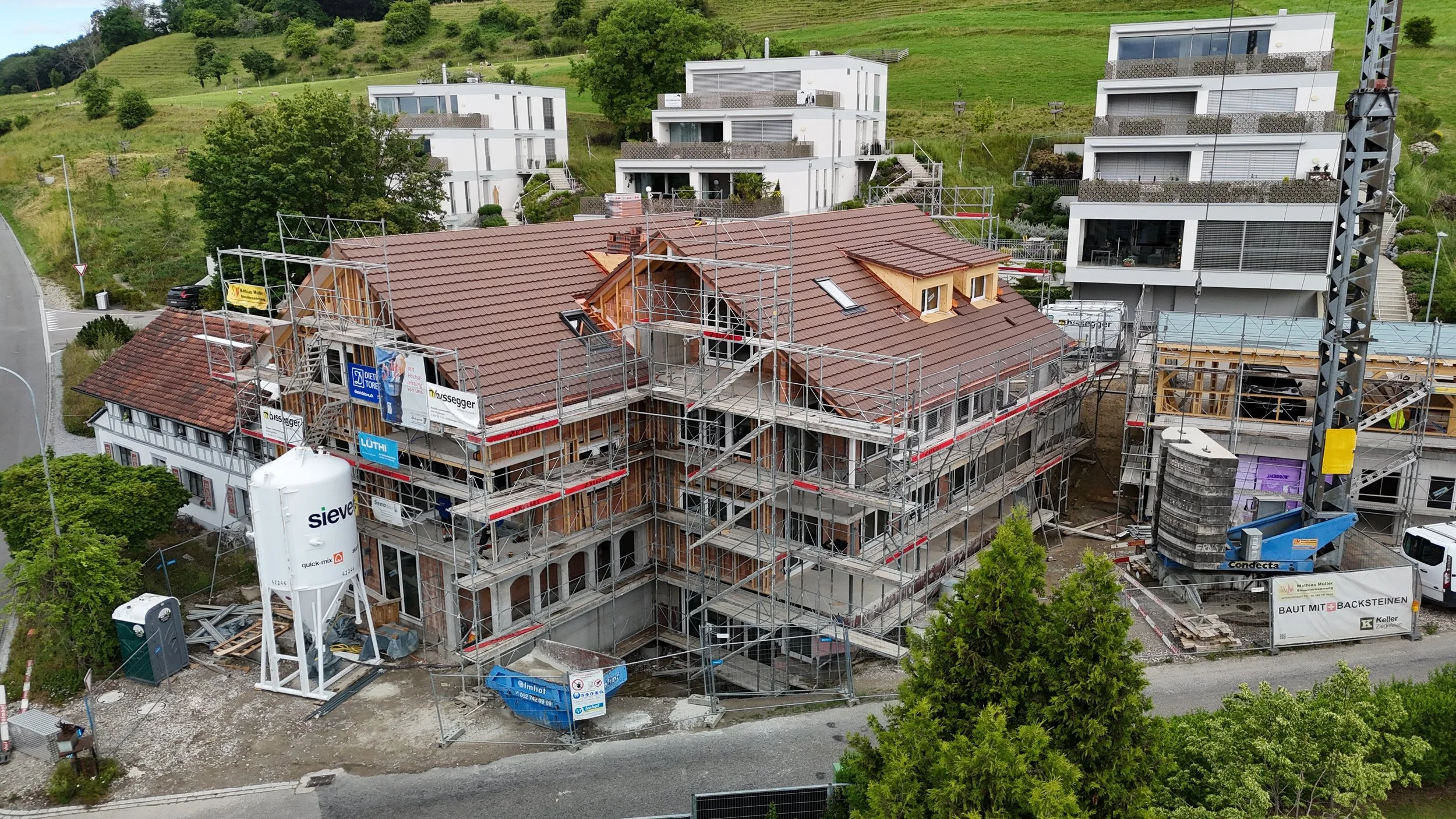 Mehrstöckiges Gebäude im Bau mit Gerüsten, Holzrahmen und Dacharbeiten, umgeben von grünen Hügeln und modernen weißen Wohnungen im Hintergrund.