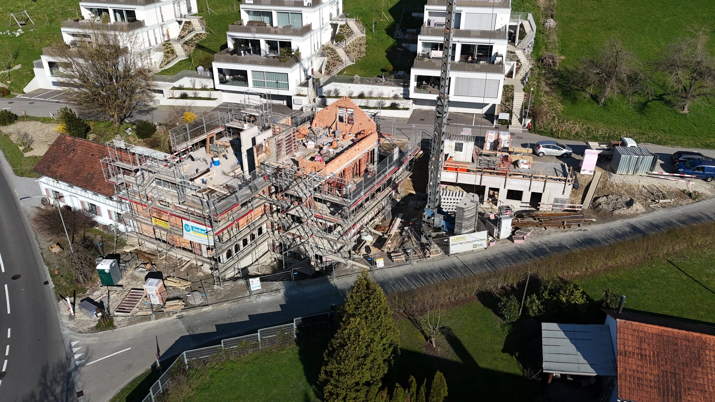 Mehrfamilienhaus im Bau, umgeben von grüner Landschaft, mit Baugerüsten und Baumaterialien auf der Baustelle.