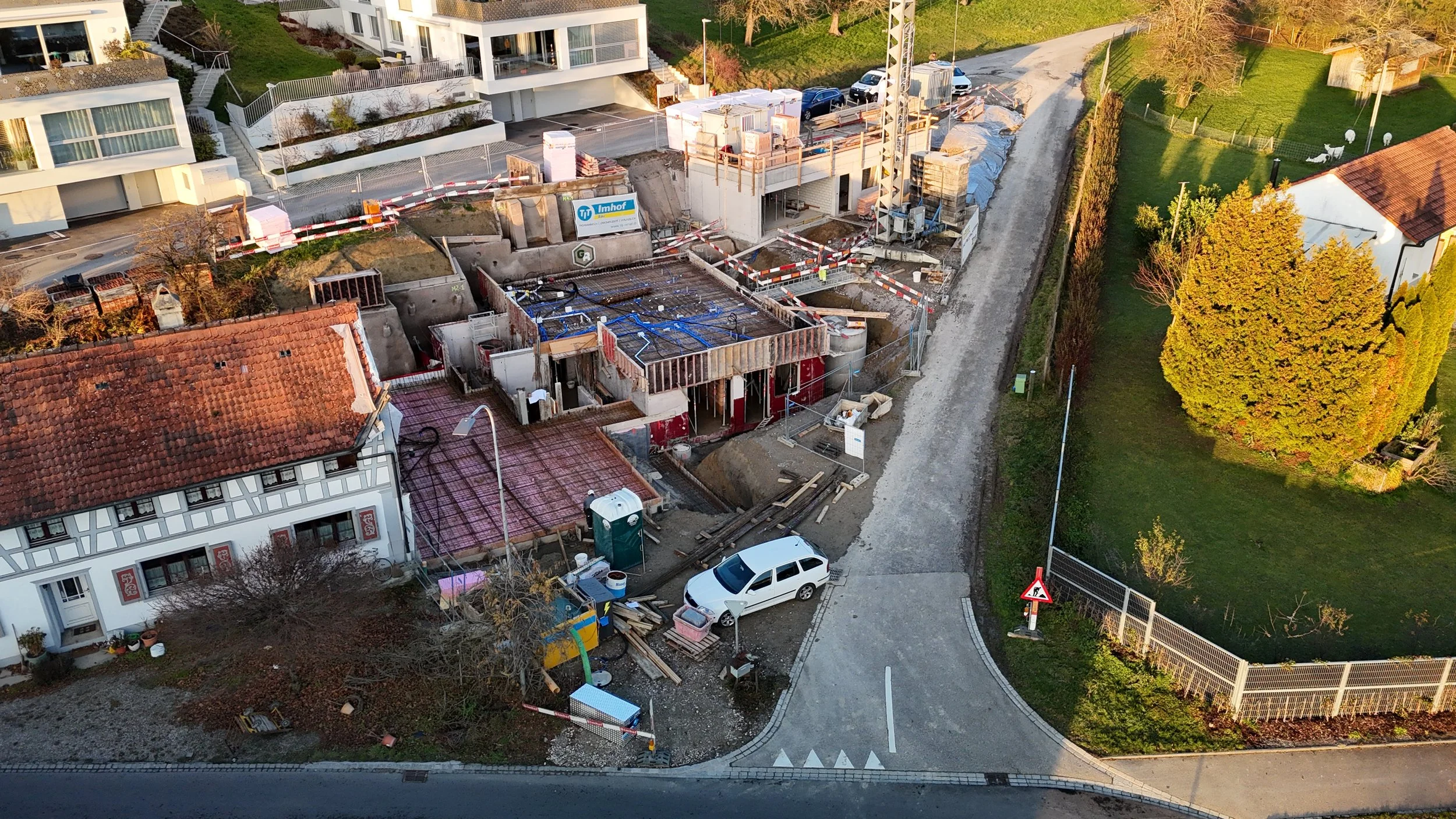 Ein im Bau befindliches Gebäude auf einer Straße, umgeben von Grünflächen und angrenzenden Wohnhäusern, mit Baustellenmaterialien und Baumaschinen.