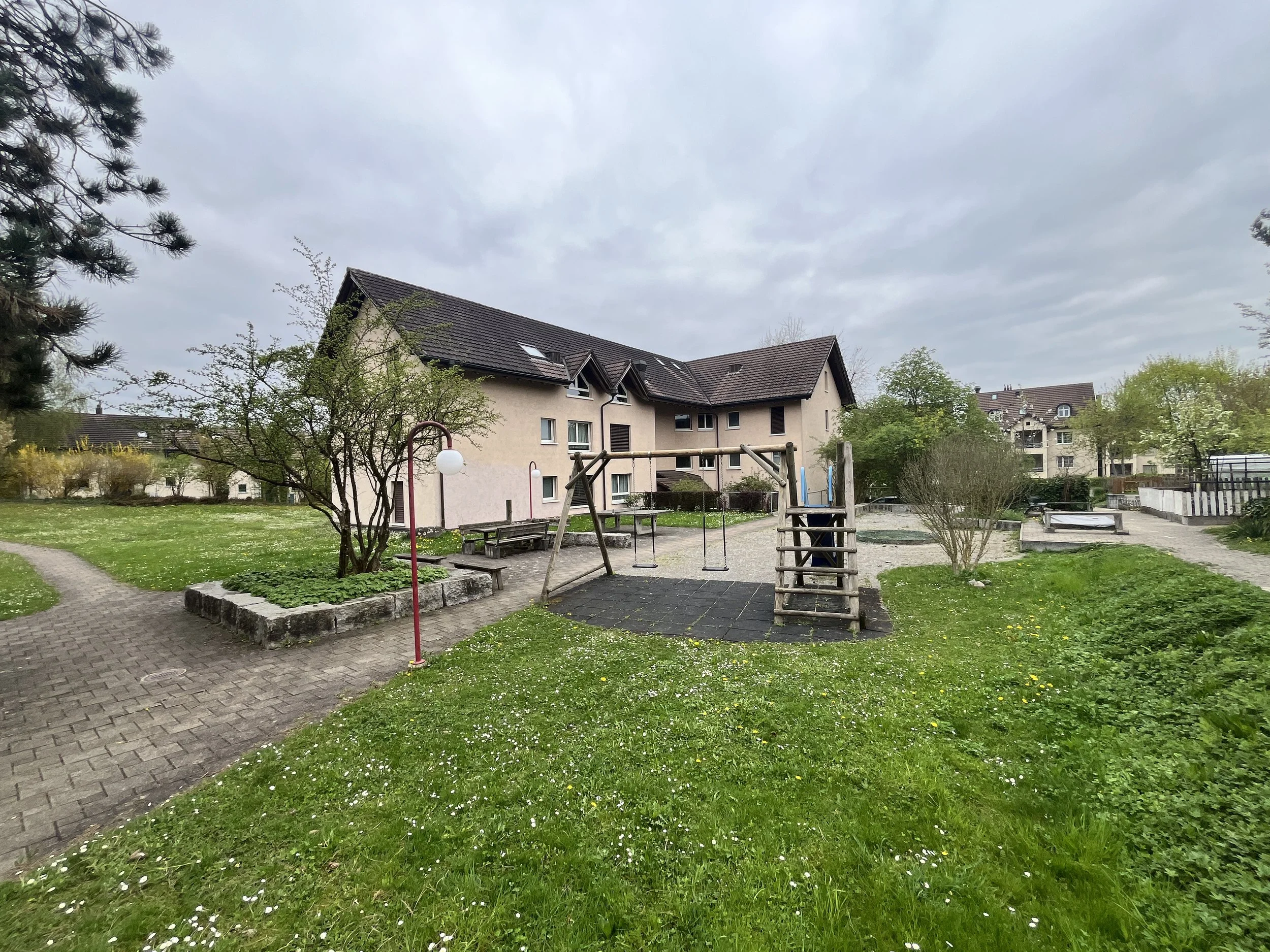 Ein Kinderspielplatz im Innenhof eines Wohnhauses, umgeben von Bäumen und grünem Rasen, bei bewölktem Himmel.