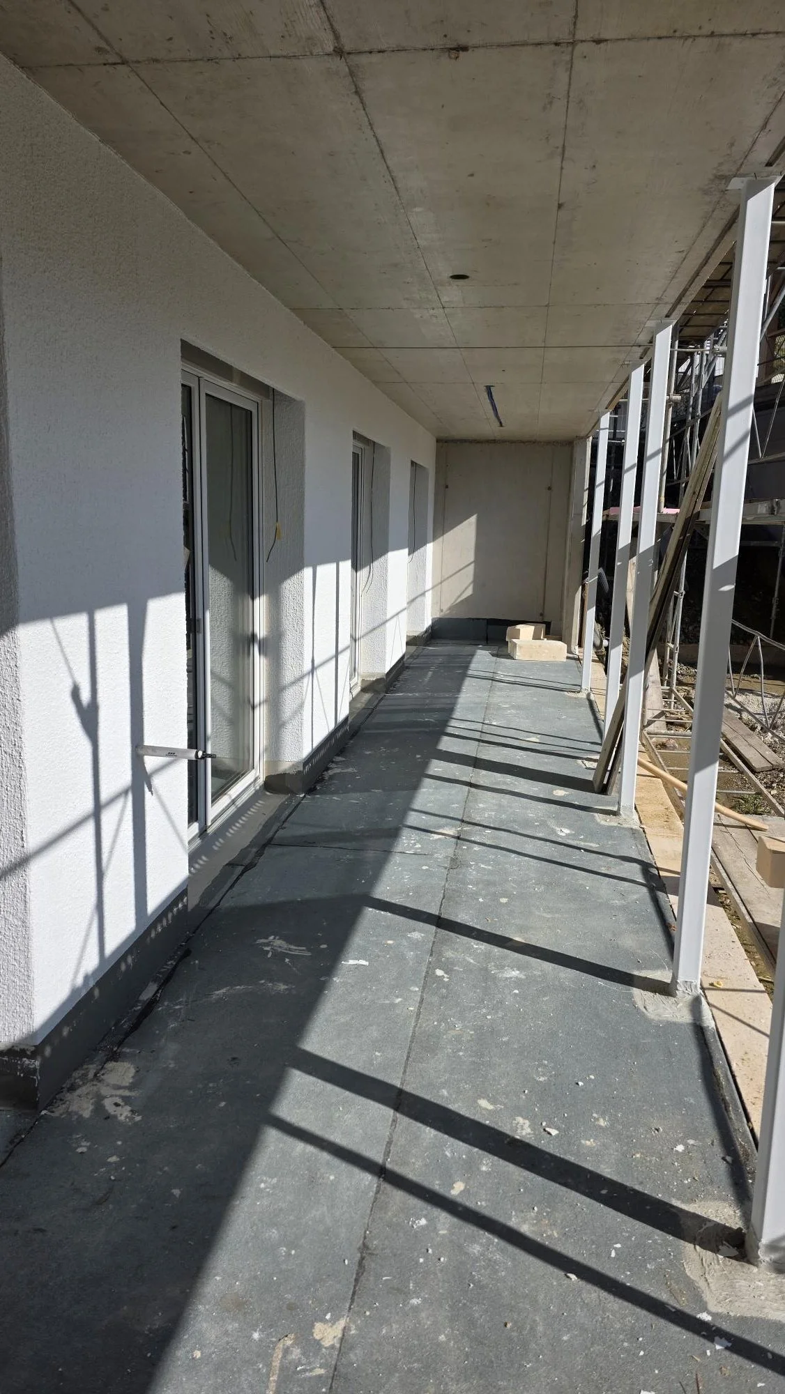 Balkon im Bau mit weißen Türen, Sichtschutzverkleidung und Baustellengerüsten, Sonnenlicht und Schatten auf der Bodenfläche.