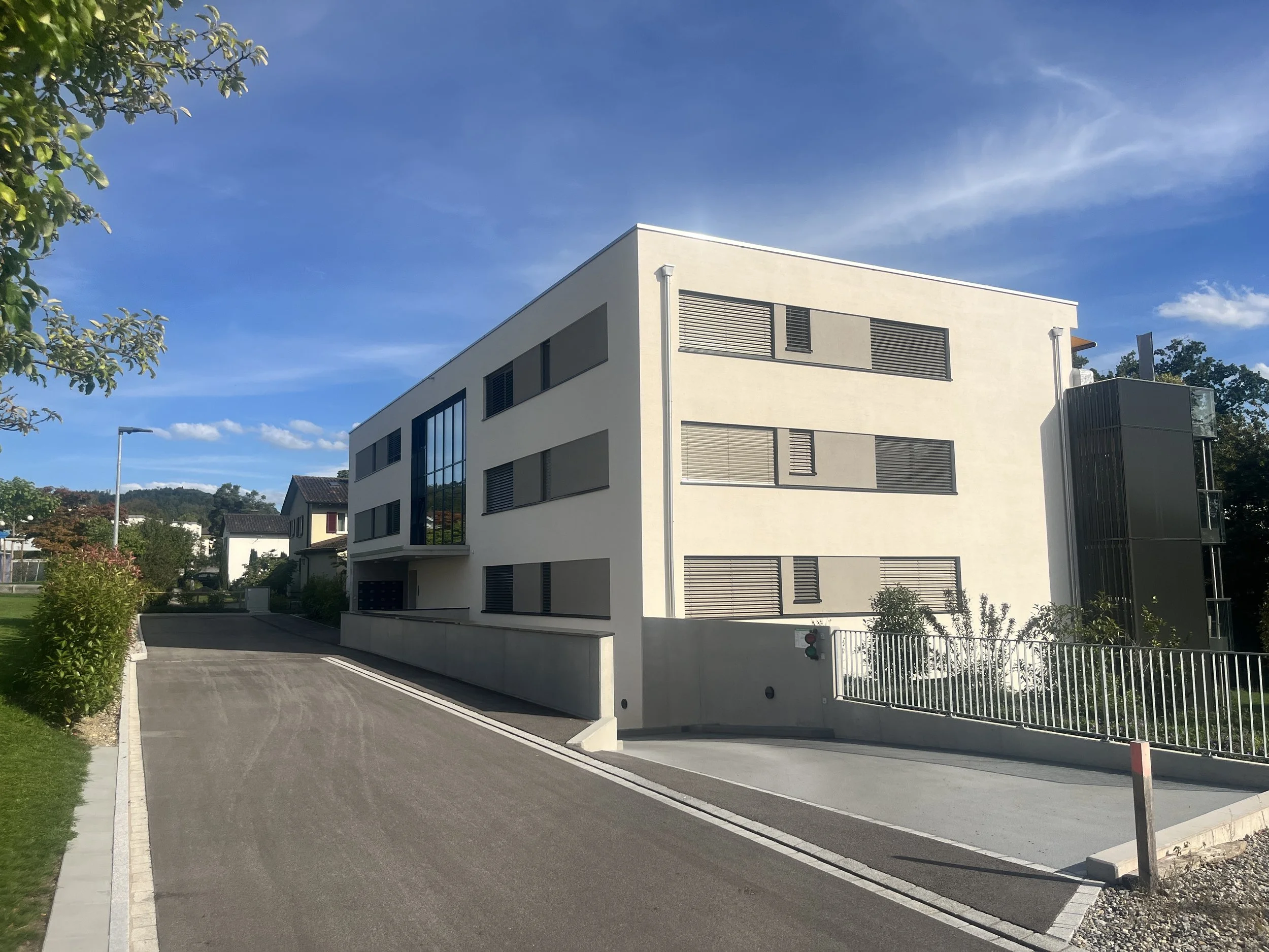 Modernes, weißes Mehrfamilienhaus mit grossen Fenstern und horizontalen Lamellen, umgeben von einem gepflegten Garten und einer asphaltierte Straße. Im Hintergrund sind weitere Häuser und Bäume sichtbar, unter einem klaren blauen Himmel.