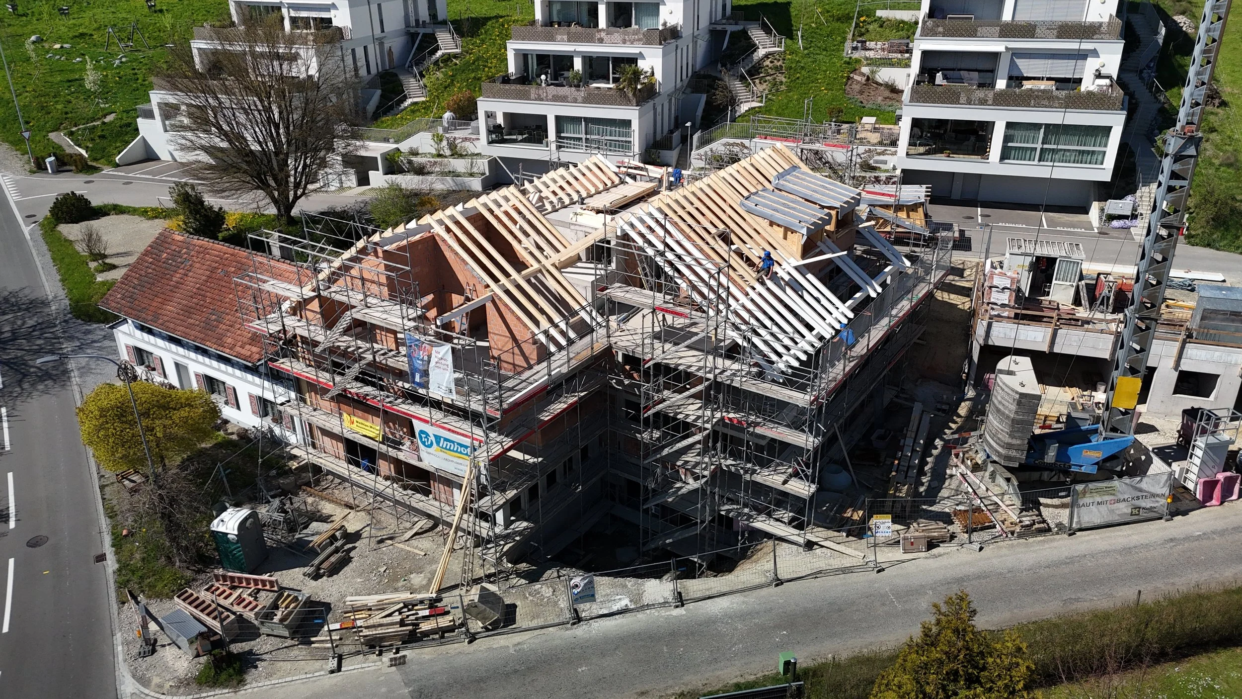 Ein Baugerüst umgibt ein Mehrfamilienhaus in Bau, das Dach wird gerade erneuert, es sind Bauarbeiten mit Arbeitern sichtbar, im Hintergrund moderne mehrstöckige Häuser in einer Wohnsiedlung.
