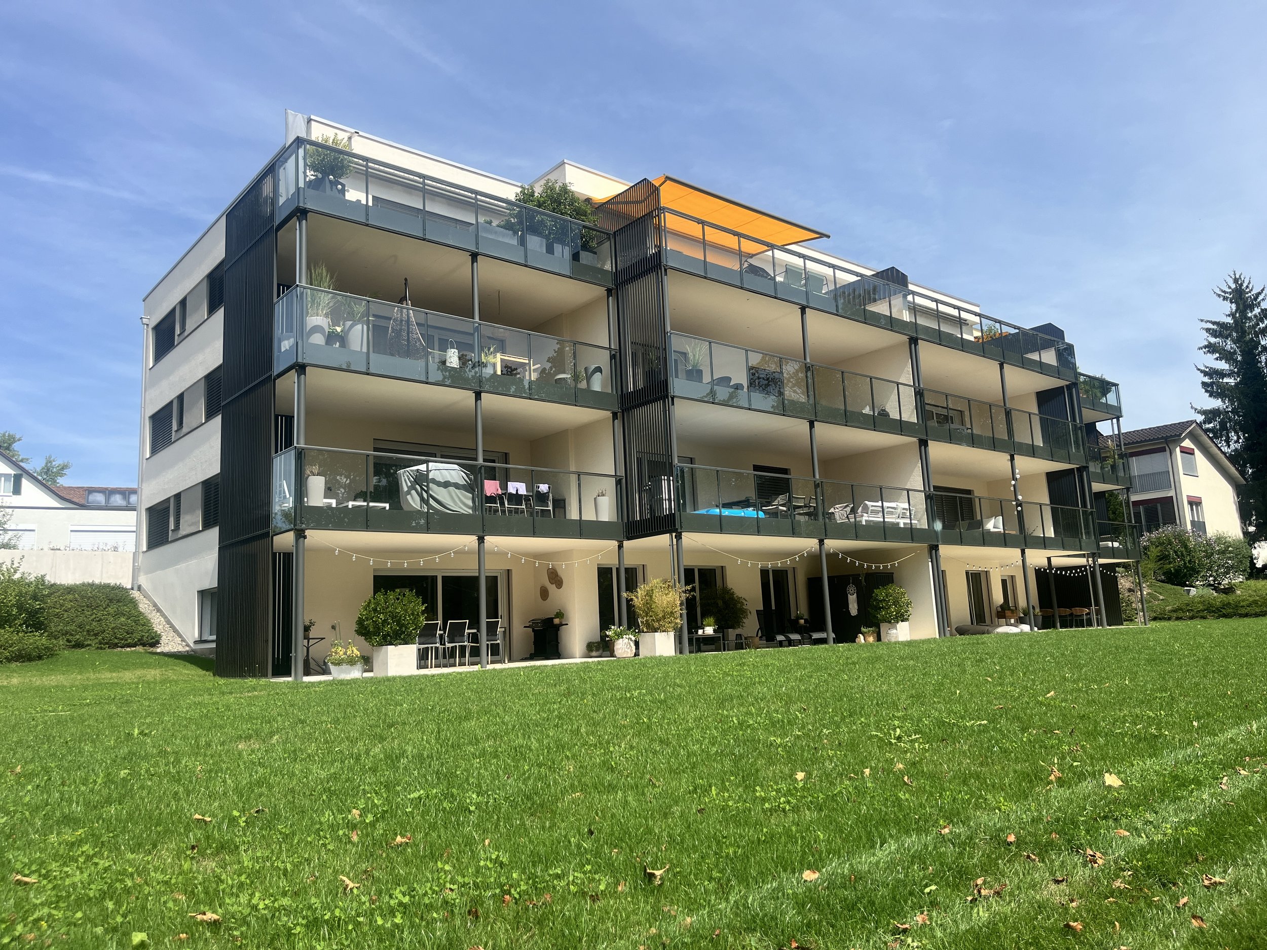 Ein modernes mehrstöckiges Apartmentgebäude mit Balkonen und Glasbrüstungen, umgeben von einem grünen Rasen und Bäumen, bei sonnigem Himmel.