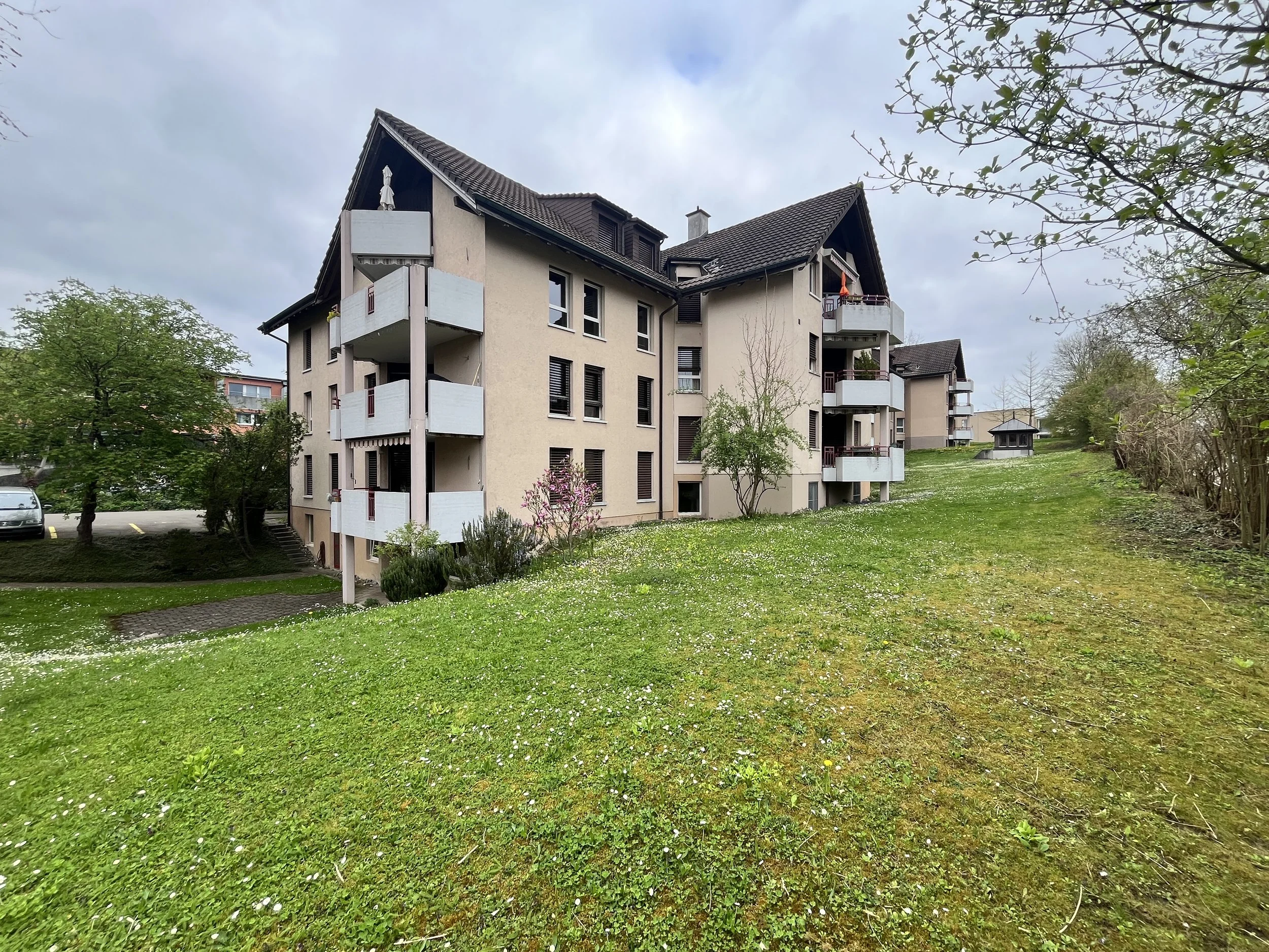 Mehrfamilienhaus in einer grünen Parkanlage mit Bäumen, Bepflanzung und Wiesen, bewölkter Himmel.