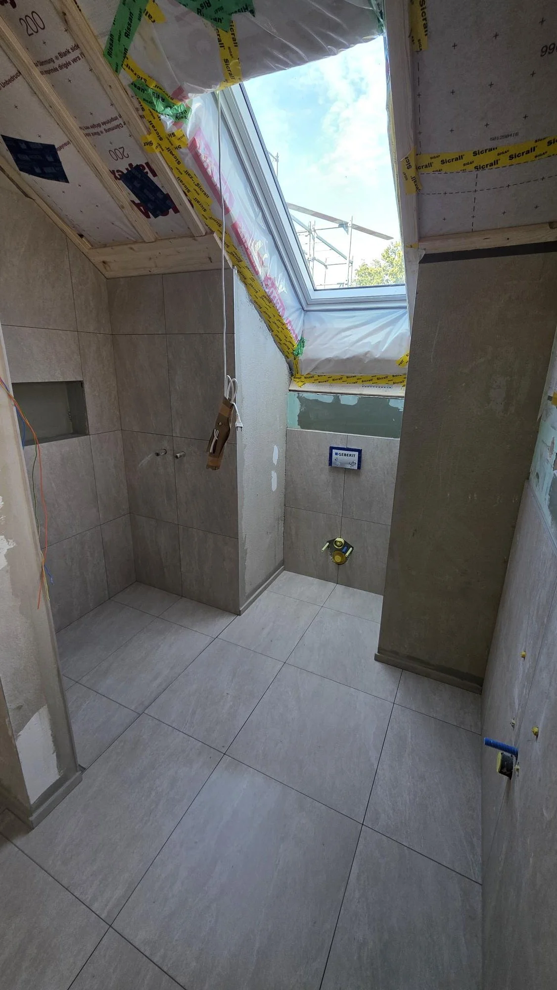 Innenraum einer eingebauten Toilette unter Bauarbeiten, mit an der Wand befestigtem Geberit-Spülkasten, Fenster mit Himmelblauem Himmel, Baustreifen um den Fensterrahmen, und unvollständig verlegte Fliesen. 