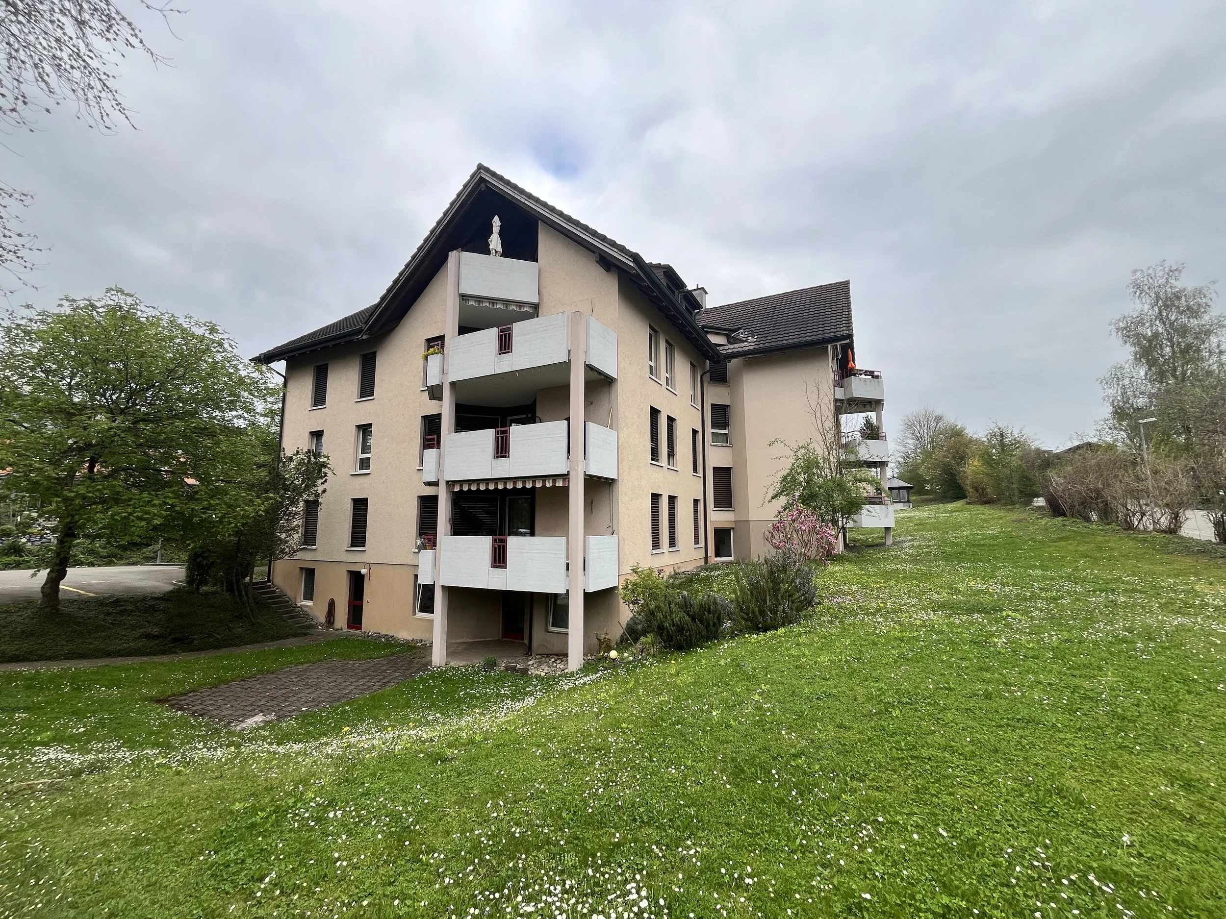 Mehrstöckiges Wohngebäude mit Balkonen vor bewölktem Himmel, umgeben von grünen Bäumen und einem blühenden Rasen.