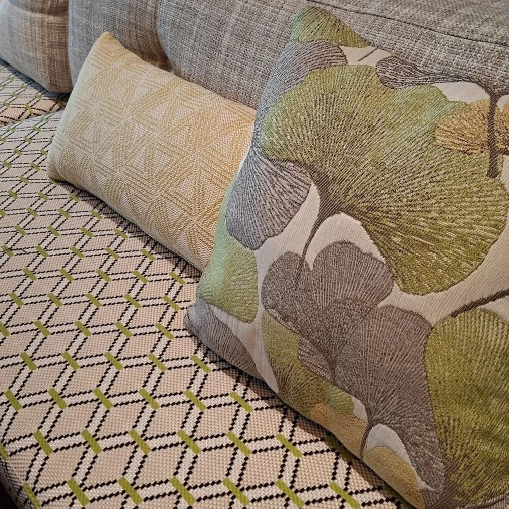Twee sierkussens op een gestreepte bank, een met een geometrisch patroon en de ander met een bladmotief in groen en bruin. Op maat gemaakt door Katrijn De Waele.