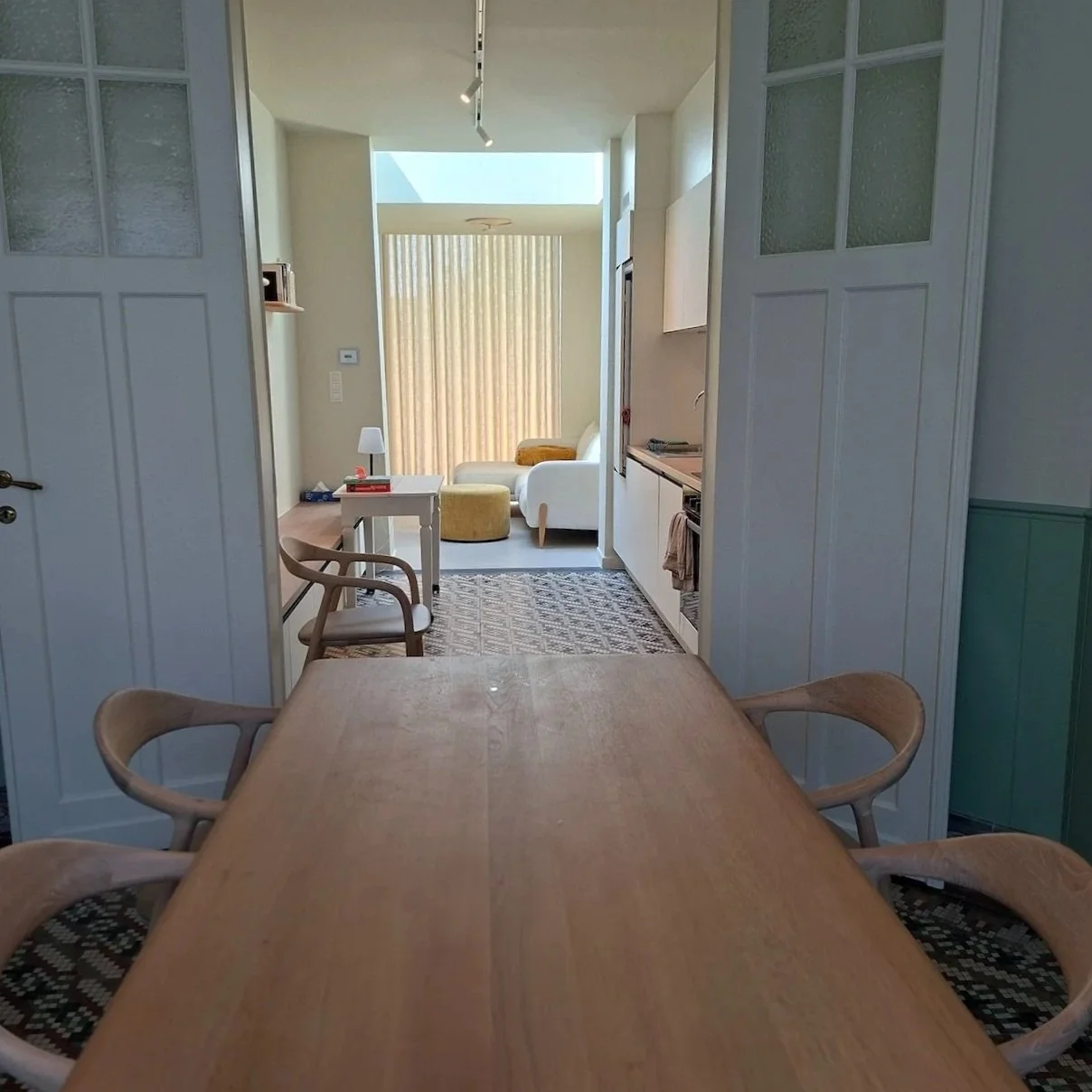 Uitzicht vanuit een eetkamer naar een woonkamer met een grote raam met gele gordijnen, een witte bank, een klein wit bijzettafeltje en een gele poef.