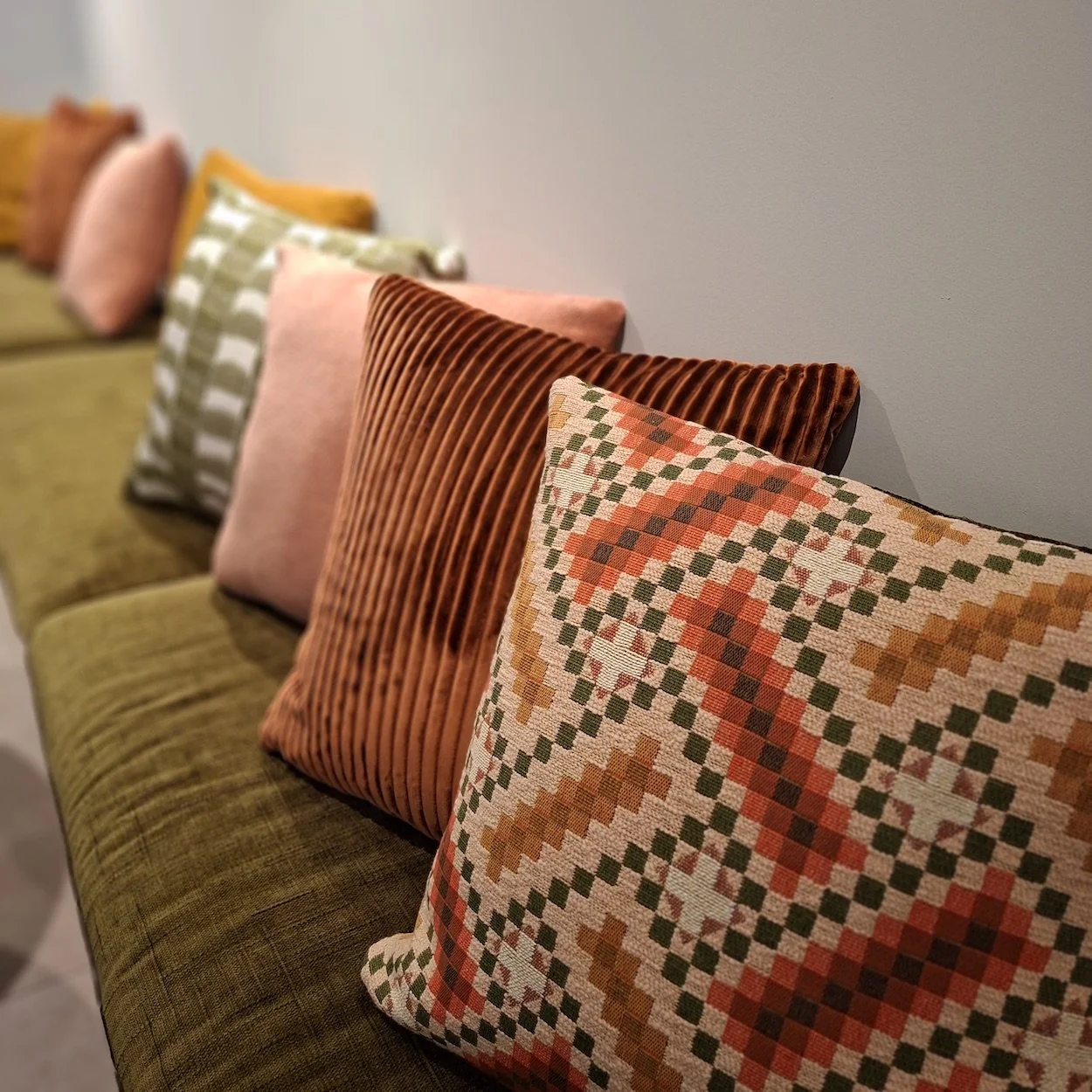 Een bank met meerdere op maat gemaakte gekleurde kussens door Katrijn De Waele. De kussens zijn in verschillende patronen, waaronder een met een geometrisch patroon in oranje, groen en beige, en de bank is bekleed met groen fluweel.