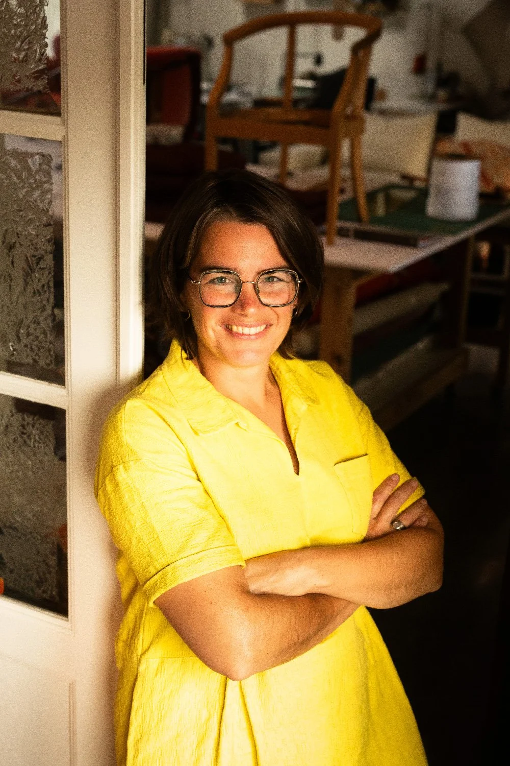 Vrouw - Katrijn De Waele - met bruin haar en glazen, lachend, gekleed in een gele jurk, staat met gekruiste armen voor haar atelier in Haacht..