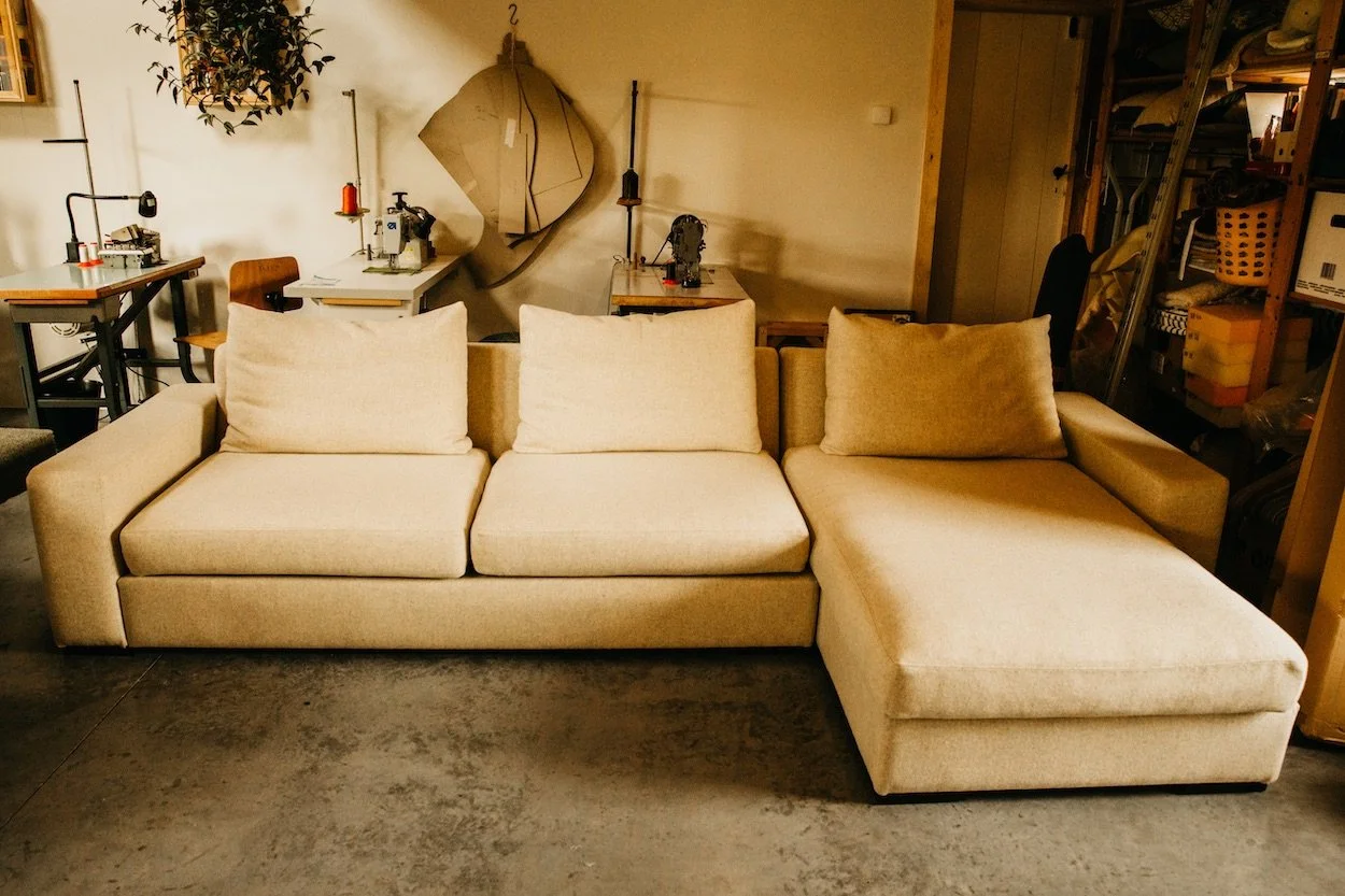 Beige 3-zitter, geherstoffeerd door Katrijn De Waele, met op de achtergrond naai- of werktafels en planken met dozen en materialen.