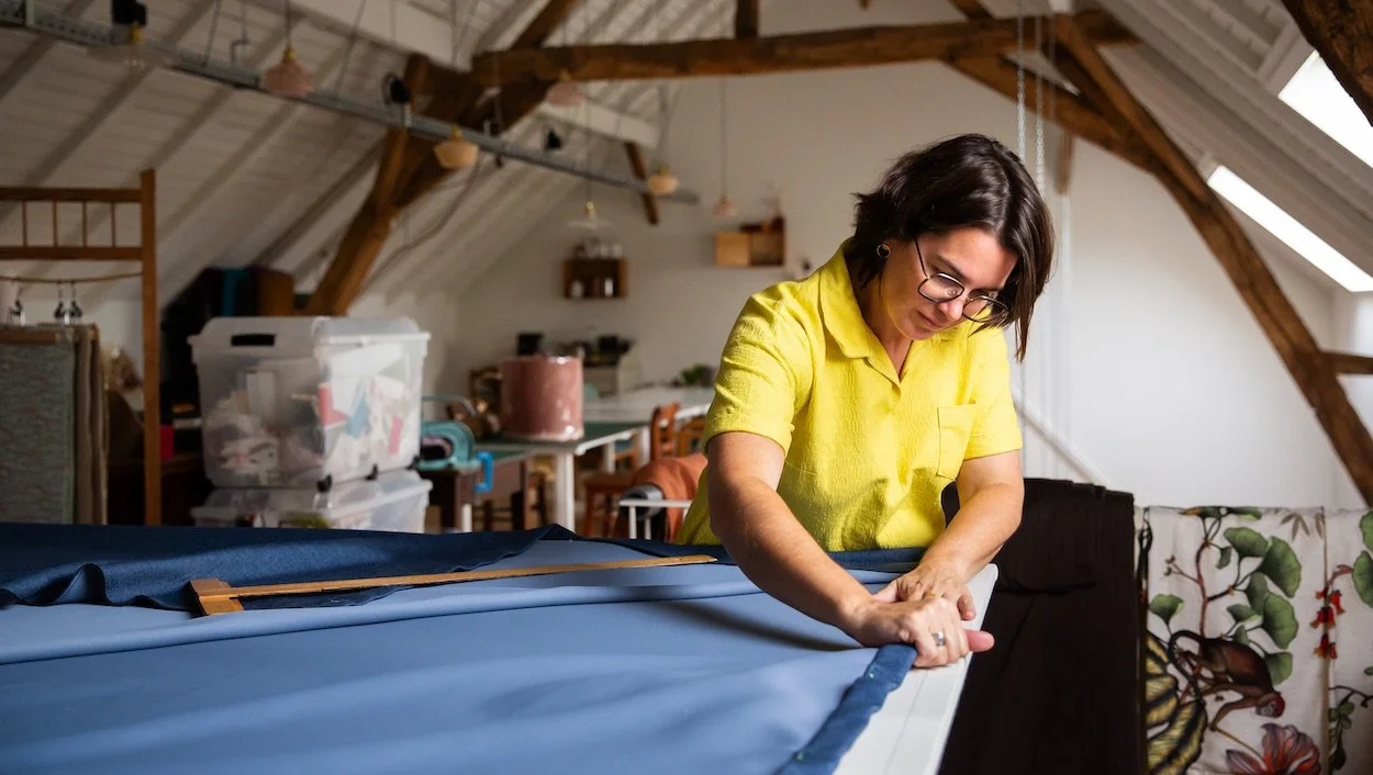 Vrouw in gele blouse - Katrijn De Waele - werkt aan gordijnen op maat in haar atelier in Haacht. Er zijn kasten, plastic opbergdozen, en planten in de achtergrond.