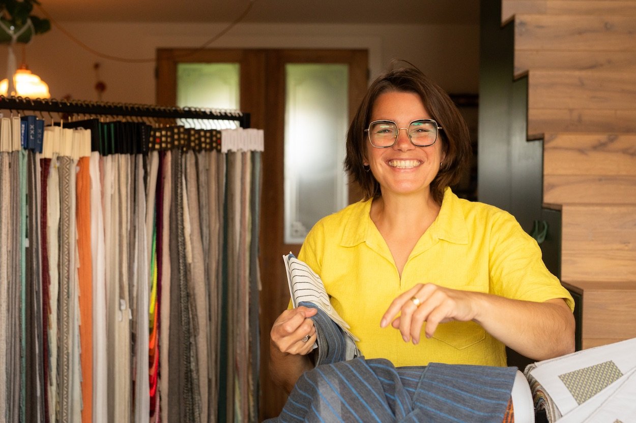 Vrouw - Katrijn De Waele - in gele blouse met grote bril, lachend met stoffen en kleurstalen in een interieur met houten en groene details.