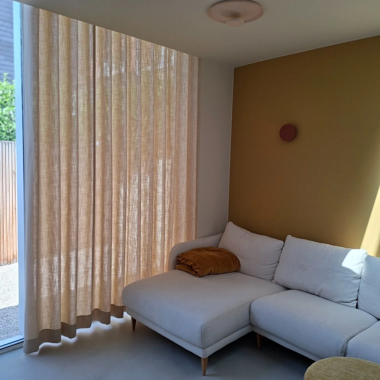 Woonkamer met een witte hoekbank, beige op maat gemaakt gordijnen door Katrijn De Waele, een gele muur met een rood decorstuk, en een plafondlamp.
