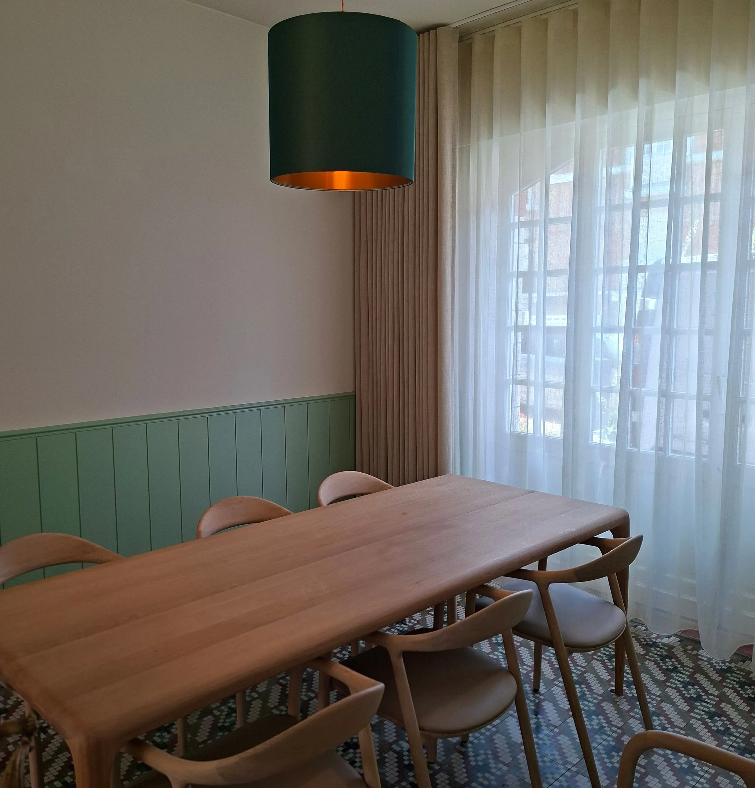 Binnenruimte met een houten eettafel en houten stoelen, muur met groene houten panelen, een grote raam met witte gordijnen en een hanglamp met groene kap.