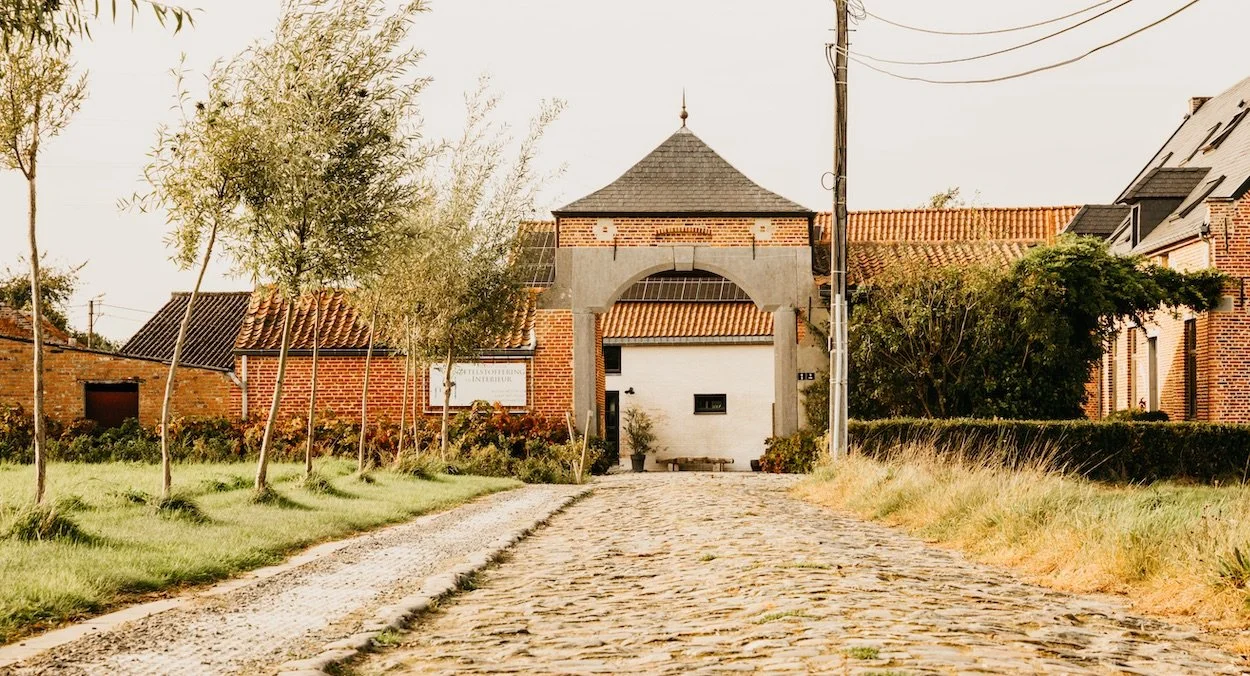 Een landweg met steentjes leidt naar een wit gebouw met een boogpoort en een beeld van een huis in een landelijke omgeving met bomen en huisjes. Dit is het atelier van Katrijn De Waele.