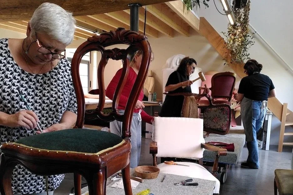 De workshop Open Atelier bij Katrijn De Waele. Vijf mensen werken aan het repareren of maken van meubels in een werkplaats, met verschillende gereedschappen en meubelstukken