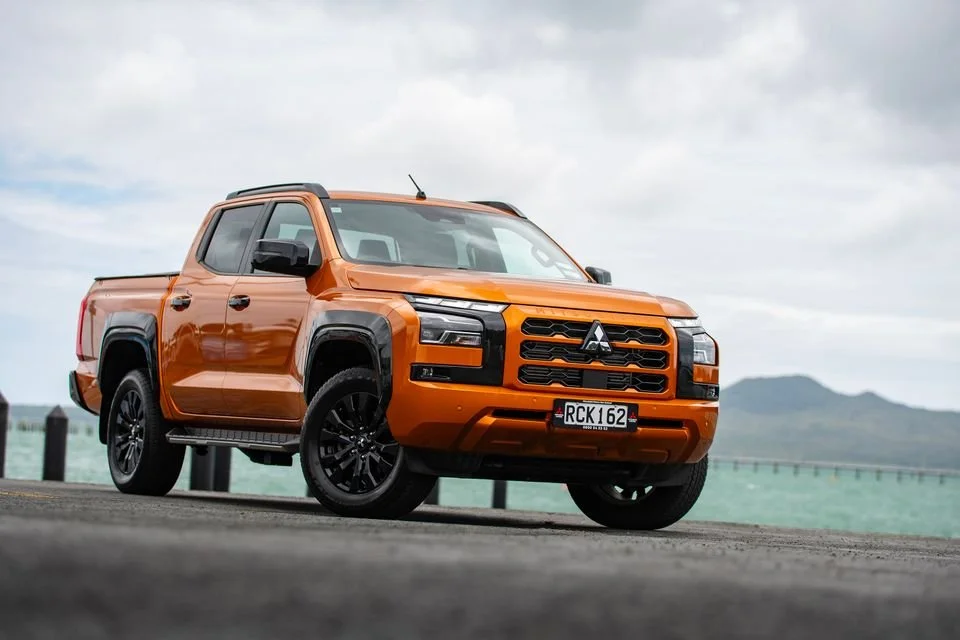 Orange pickup truck parked on the side of a paved road near water, with mountains and cloudy sky in the background.