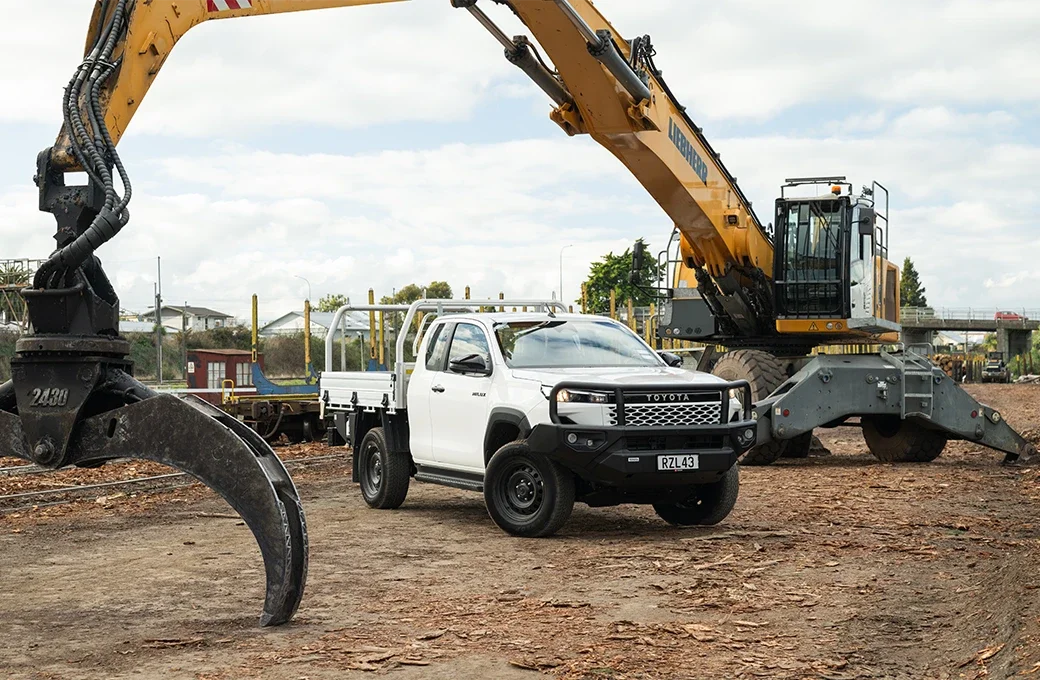 A construction site with a large yellow excavator next to a white pickup truck, both on dirt ground, under a partly cloudy sky.