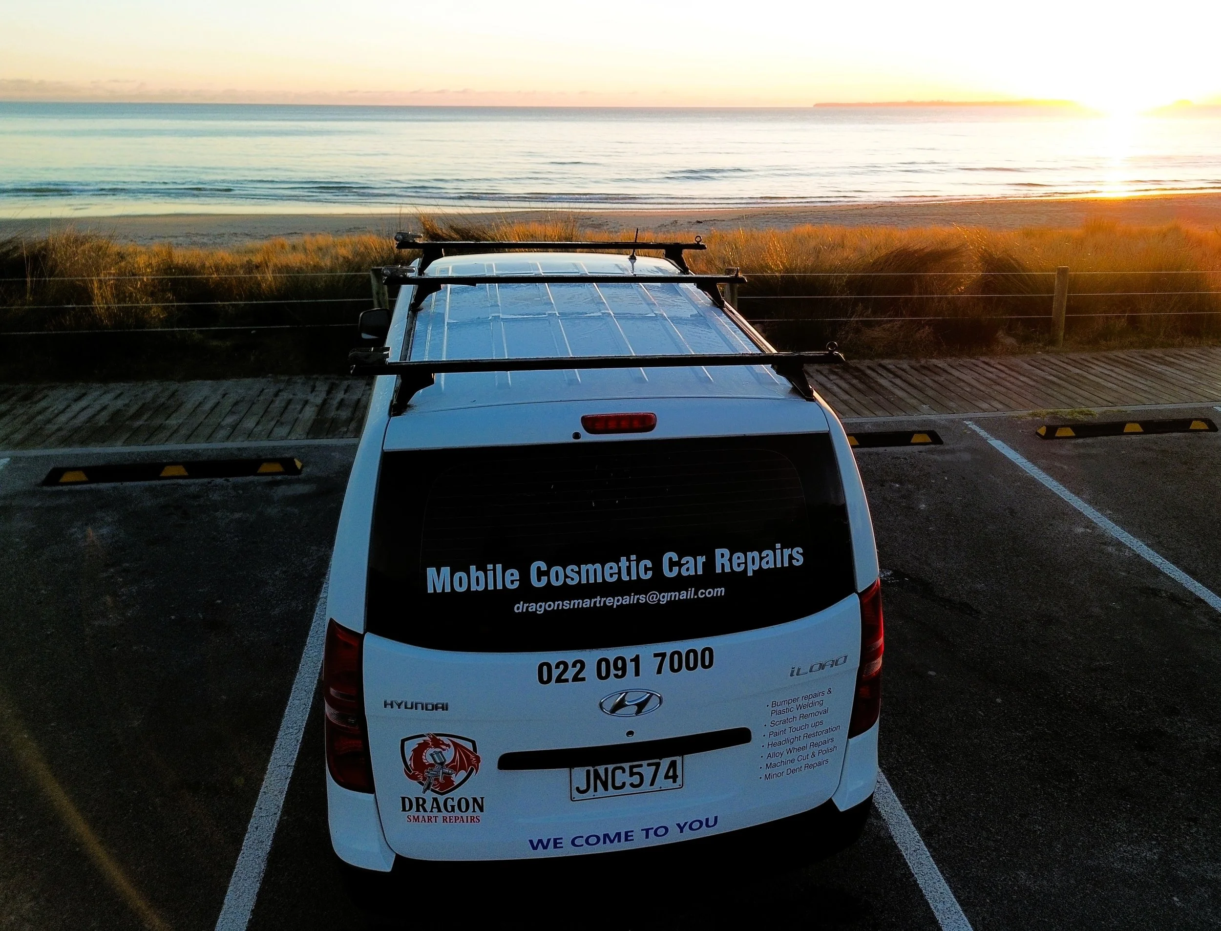 A white Hyundai car parked in a parking lot near a beach during sunset, with a sign advertising mobile cosmetic car repairs on the back of the car.