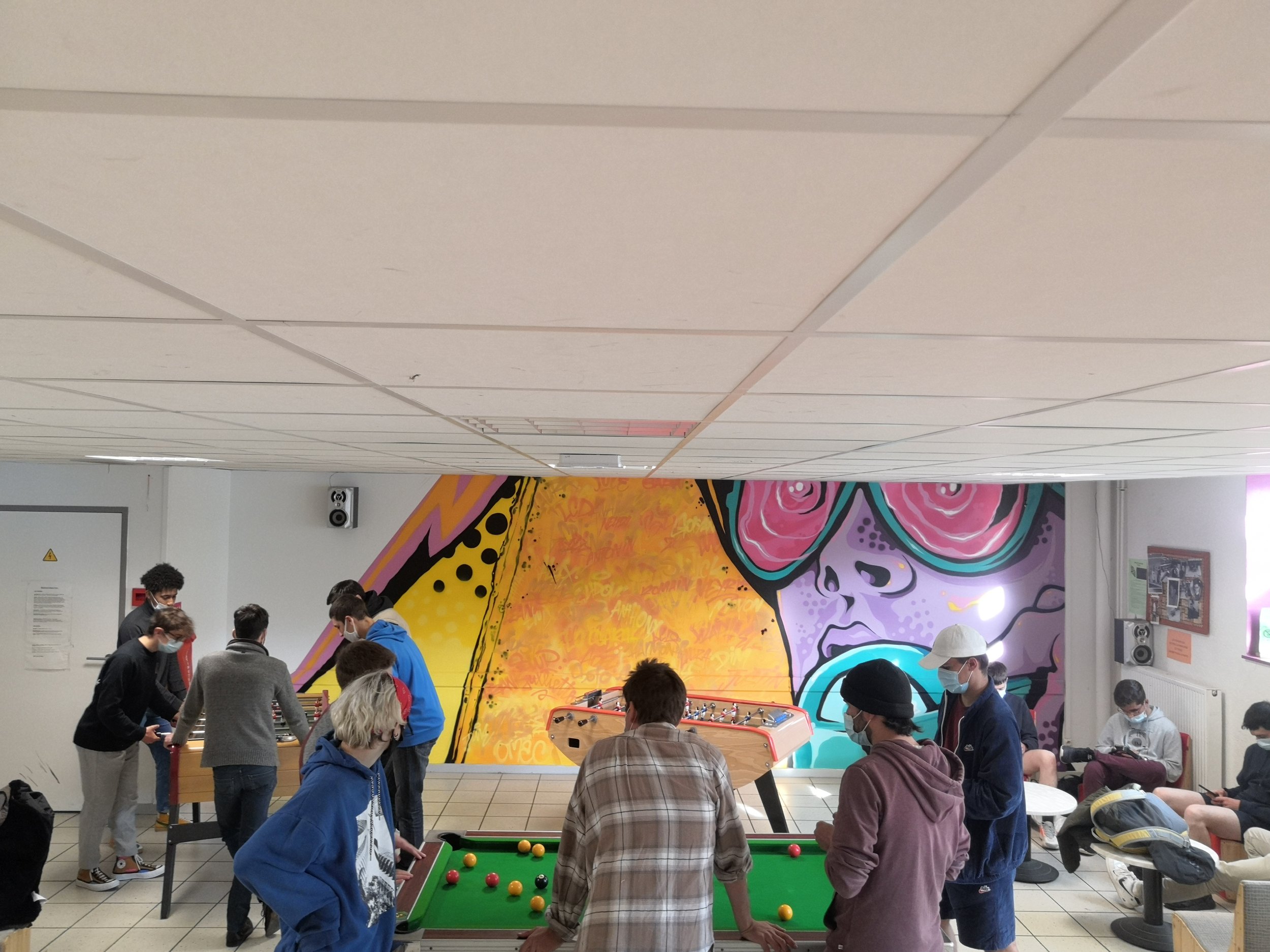 Groupe de personnes jouant au baby-foot dans une salle d'une exposition ou d'une salle de loisirs, avec une grande fresque colorée représentant un squelette de calavera dans le fond.